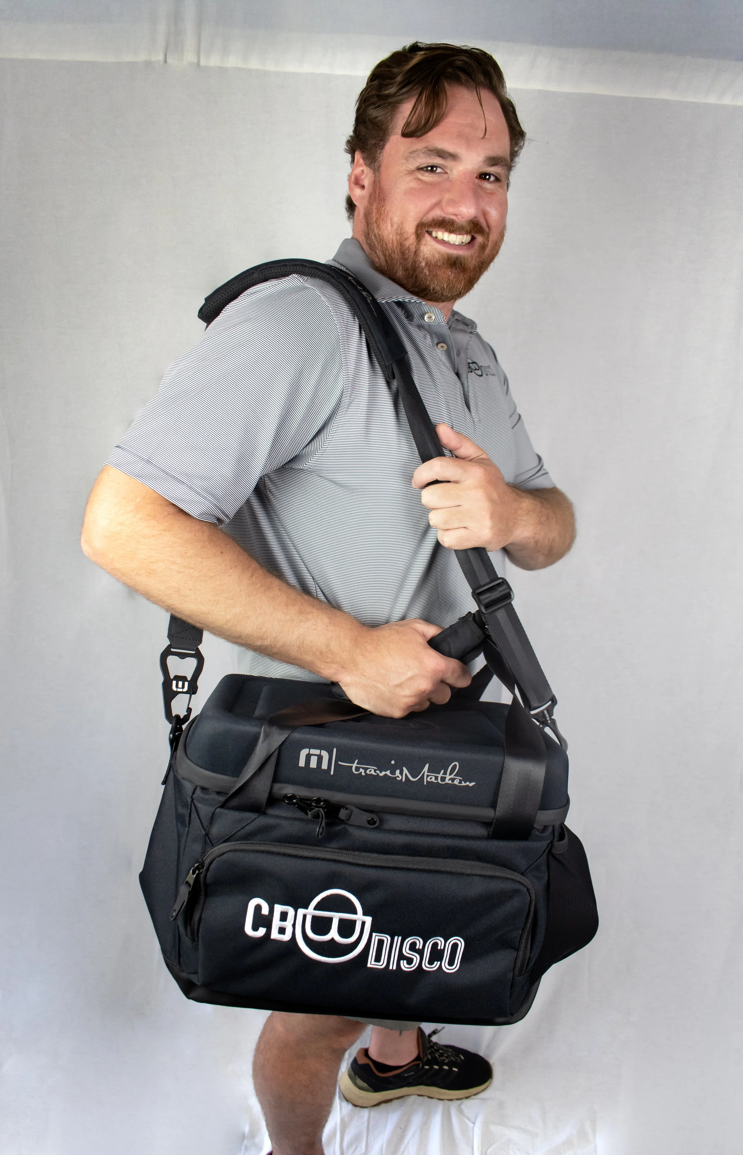 Man with a beard smiling, carrying a black travel bag with the logos 'Travis Mathew' and 'CBT Disco', standing against a plain background.
