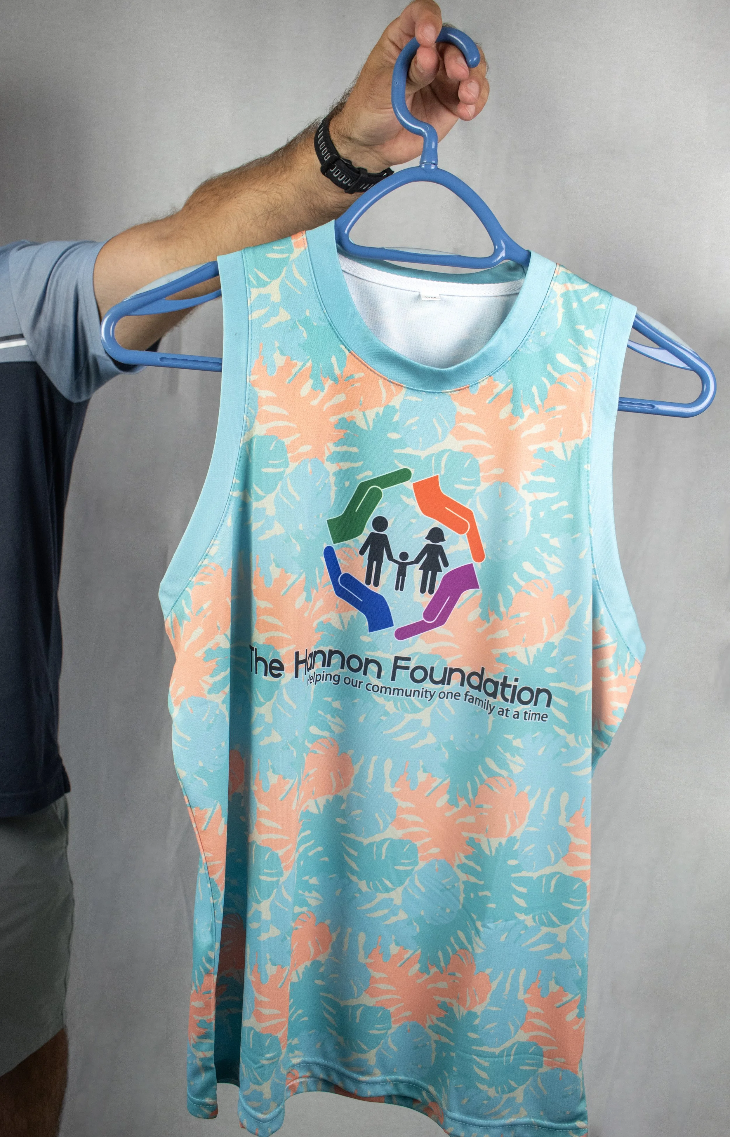 A sleeveless shirt with a tropical leaf pattern in pastel colors, featuring a logo of multi-colored hands forming a circle around a family, with the text "The Hannon Foundation helping our community one family at a time."