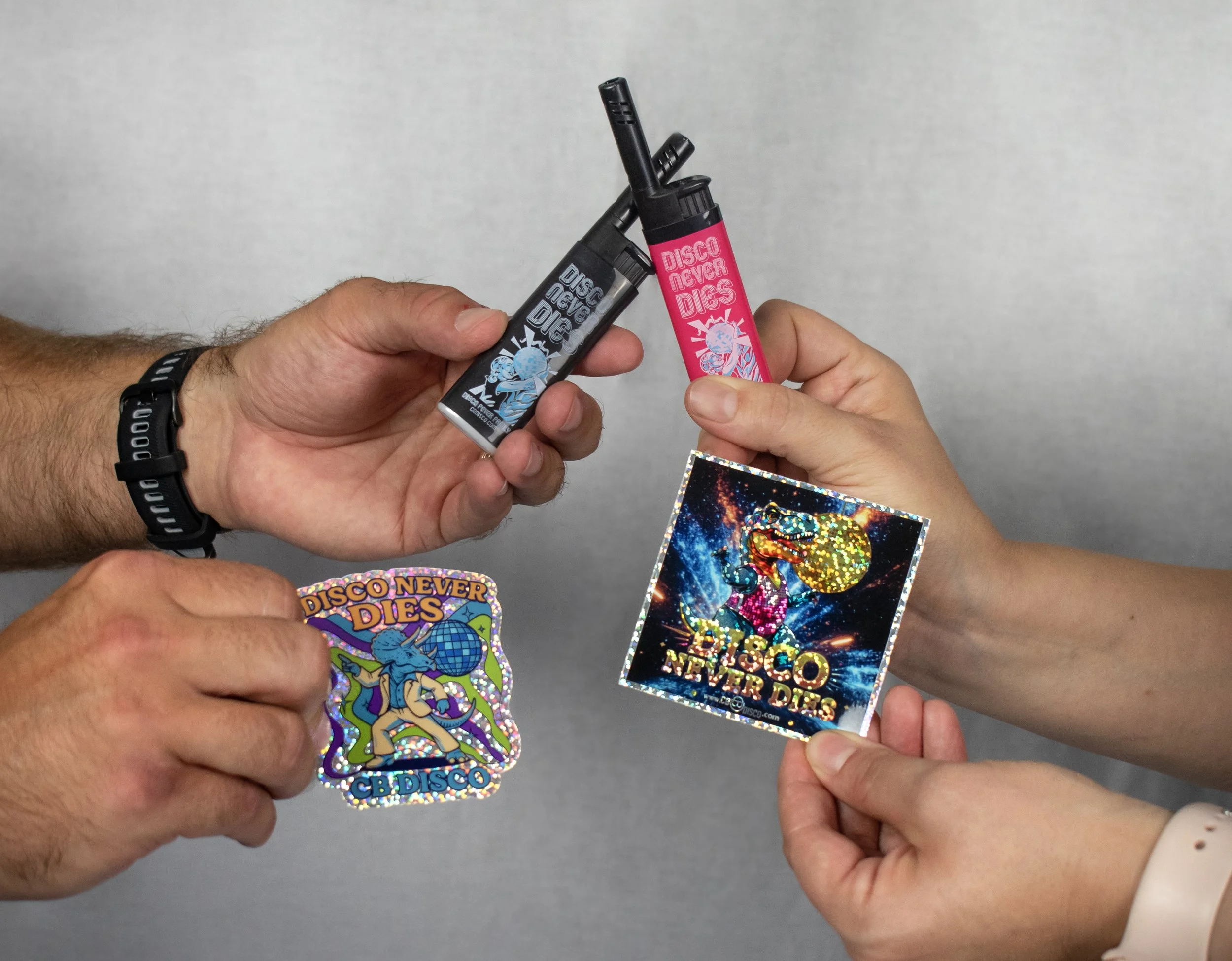 Two people exchanging colorful stickers and lighters with 'Disco Never Dies' design, against a neutral background.