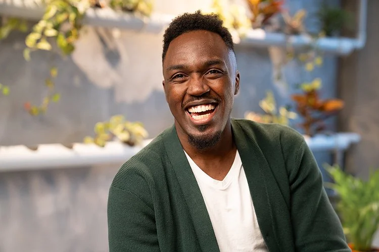 A smiling African American man with short curly hair, wearing a white t-shirt and green cardigan, sitting outdoors with a blurred background of plants and a concrete wall.