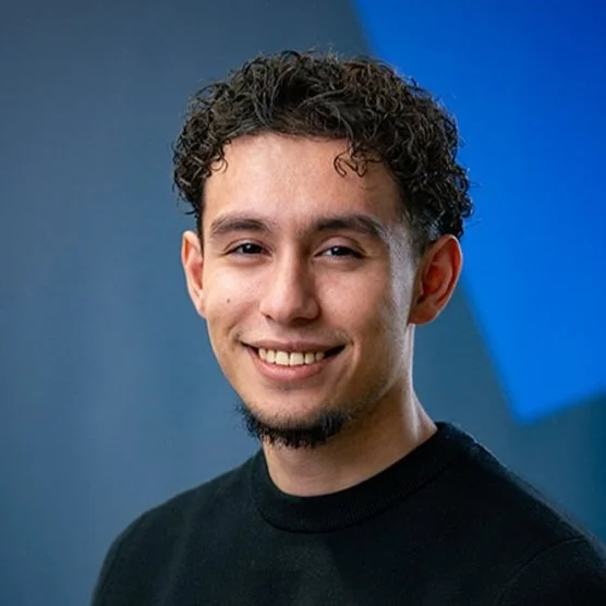 Smiling young man with curly hair and a goatee wearing a black shirt against a blue background.