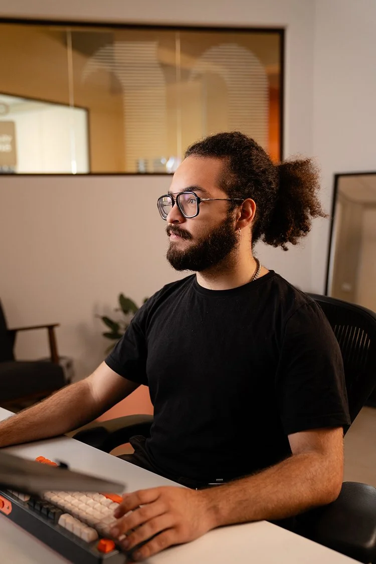 Een man met krullend haar, een bril en een baard past studioverlichting aan in een fotostudio met planten en een stoel op de achtergrond.