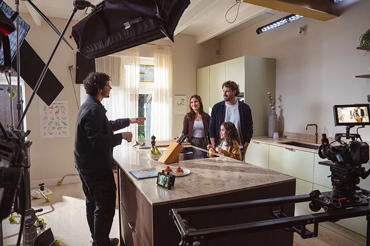 A family of four, including a man, woman, and two children, stands in a kitchen set with a man in a black hoodie directing them, during a video shoot with professional camera and lighting equipment.