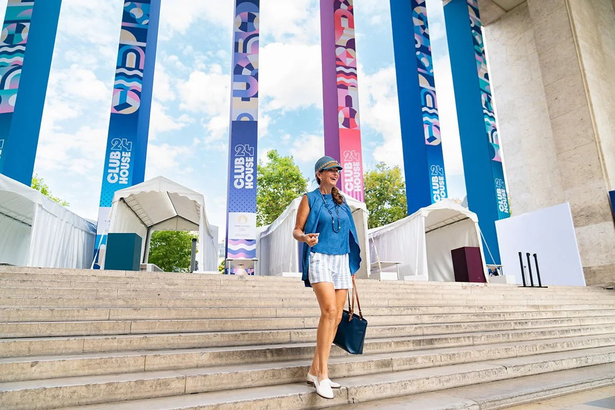 A woman in summer clothing standing on outdoor steps in front of colorful banners with abstract designs, reading 'CLUB 24 HOUSE.' The sky is partly cloudy with some trees visible in the background.