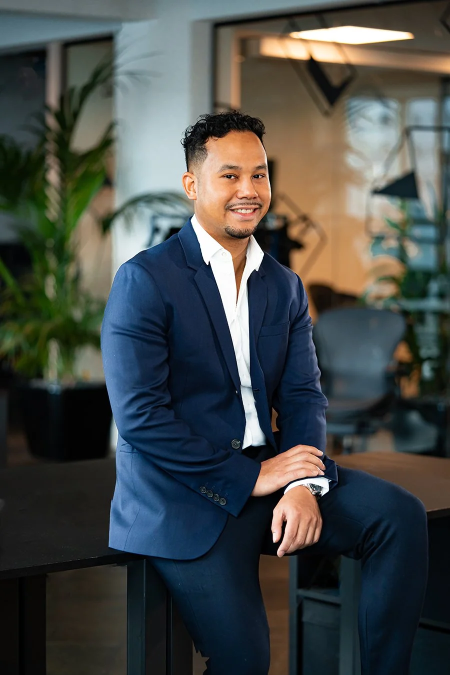 A man in a navy blue suit and white shirt sitting on a black table in a modern office, smiling at the camera with plants and office furniture in the background.