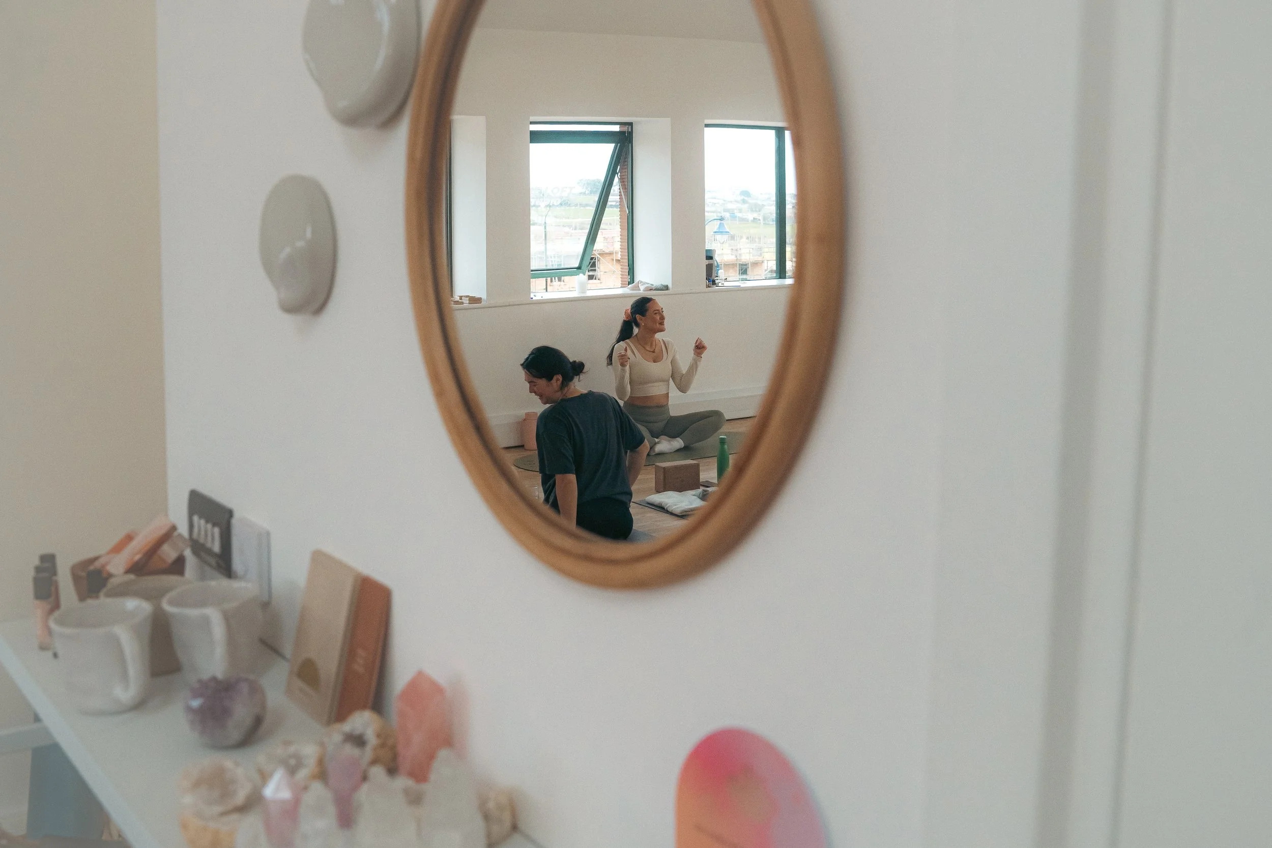 Reflection of two women practicing yoga in a room with large windows, one sitting cross-legged and the other kneeling with arms raised.