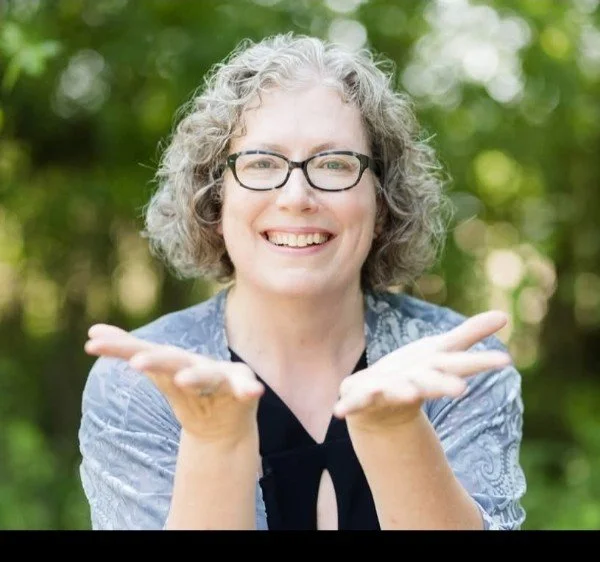 A smiling woman with glasses and curly gray hair outdoors, holding her hands out in front of her with palms up.