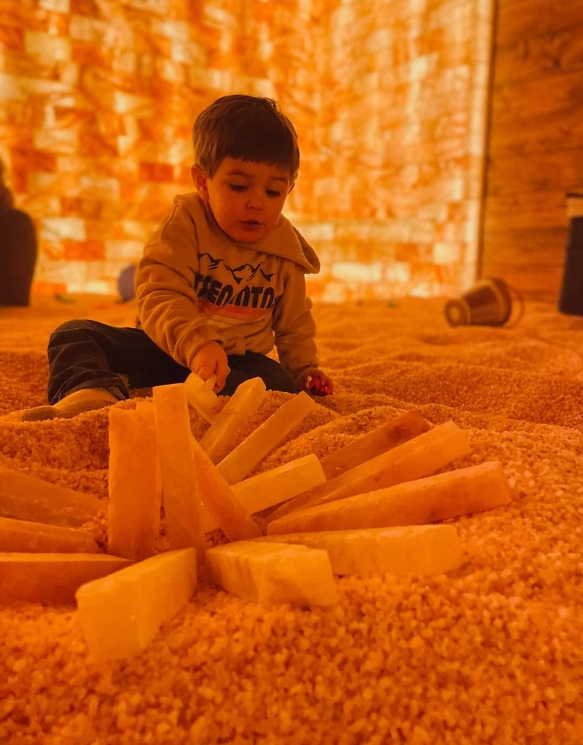 Child in a yellow jacket playing with wooden blocks on a sandy surface inside a warmly lit room.