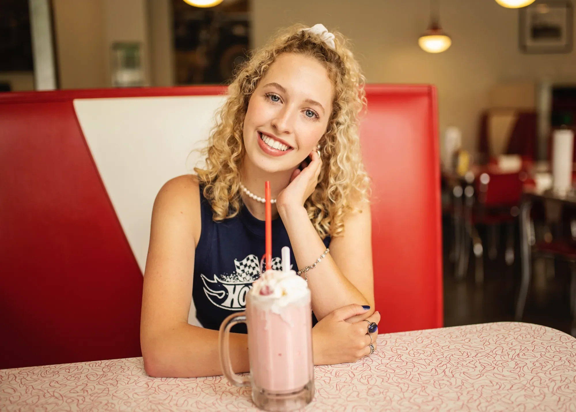 A young woman with curly blonde hair smiling in a diner, sitting at a pink and white table with a milkshake topped with whipped cream and a red straw in front of her.