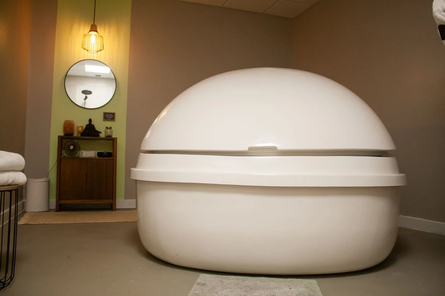 Indoor space with a large, rounded, white pod-like structure in the foreground. In the background, there is a small wooden cabinet with decorative items, a mirror on the wall, and warm lighting.
