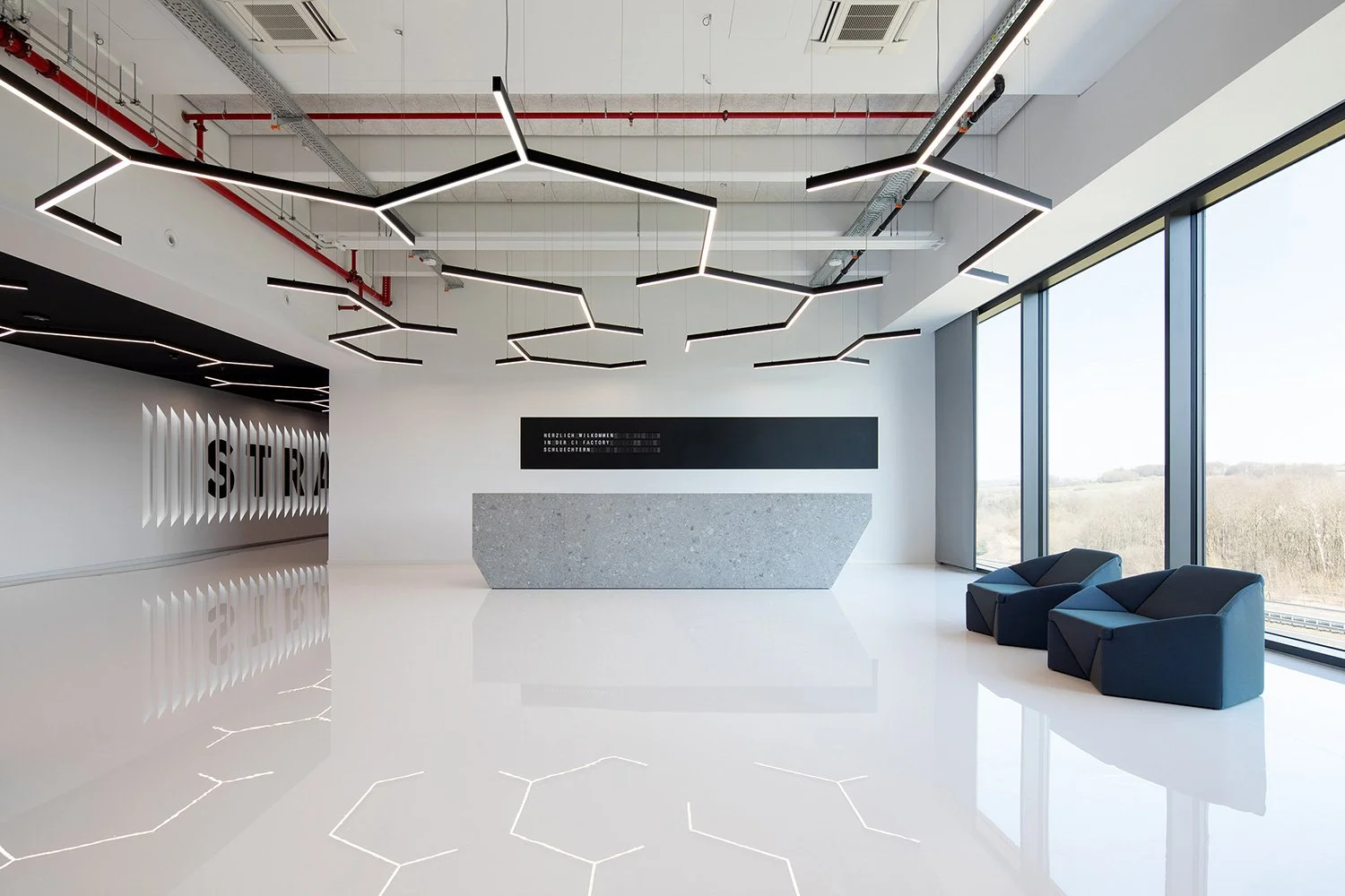 Interieuraufnahme der modernen Lobby der Engelbert Strauss CI Factory mit skulpturalem Empfangstresen, geometrischen Lichtinstallationen und angedeuteter CI-Lamellenwand.
