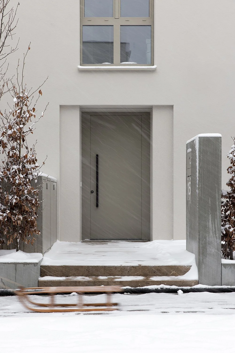 Detailaufnahme des zurückgesetzten Eingangsbereichs eines Wohnhauses am Schubertschen Park mit monochromer Fassadengestaltung, Schneefall und vorbeiziehendem Holzschlitten im Vordergrund.
