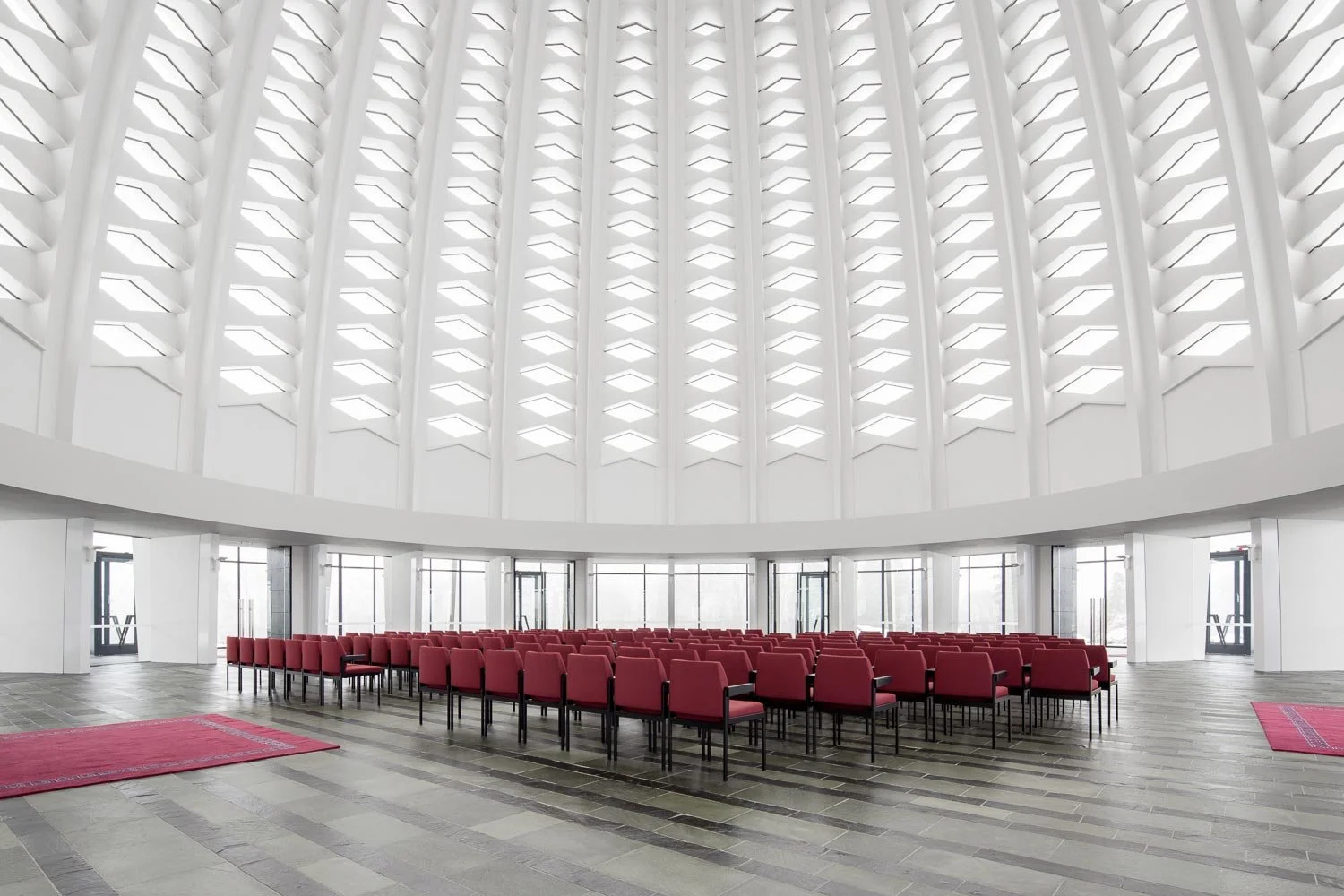 Zentralperspektive des Innenraums im Bahá'í-Tempel. Die helle, lichtdurchflutete Atmosphäre der riesigen, freitragenden Kuppel kontrastiert mit den geordneten Reihen der roten Bestuhlung.