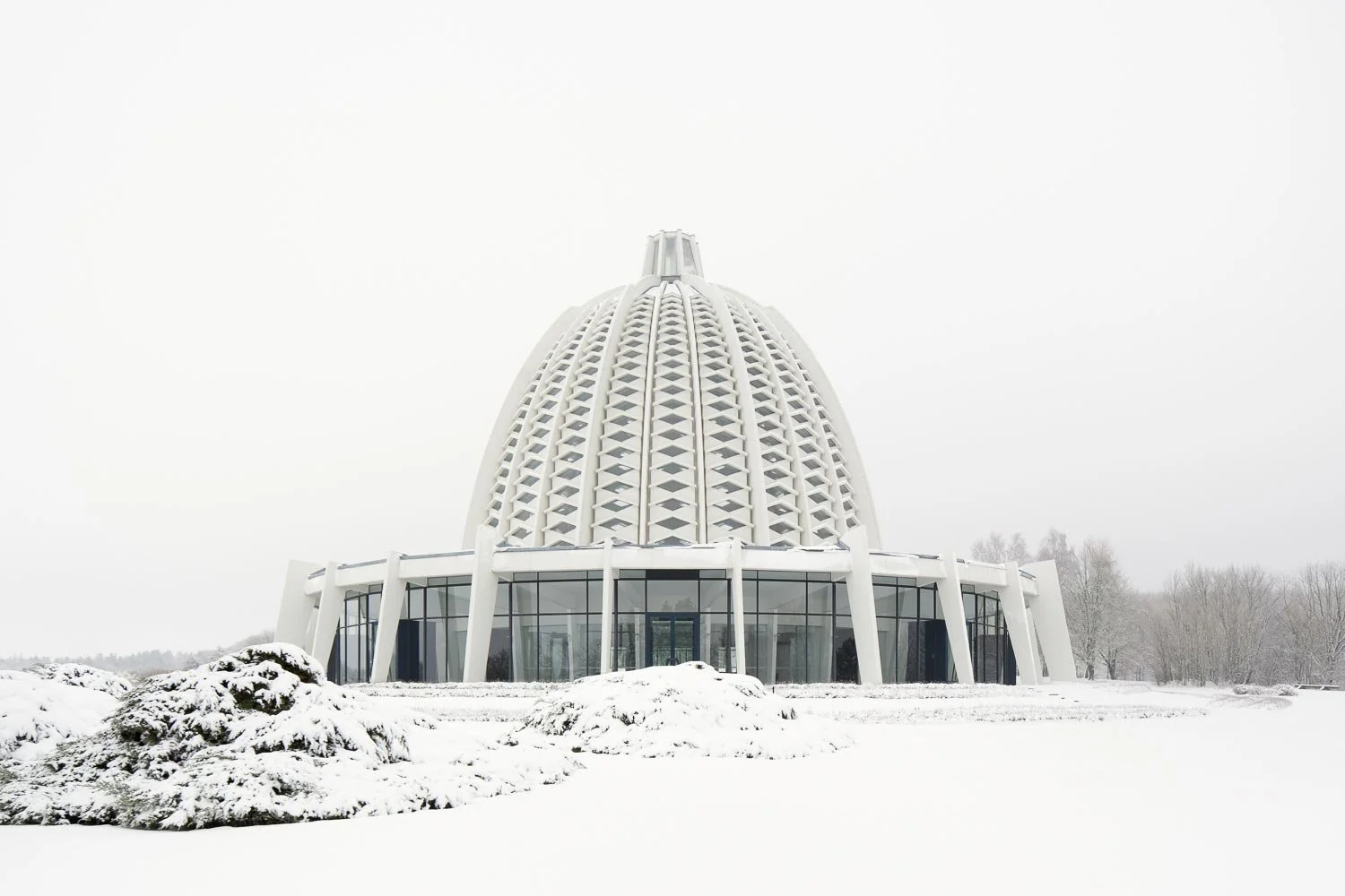 Zentralperspektive des europäischen Bahá'í-Tempels vor hellgrauem Himmel. Die lichtdurchlässige Kuppelarchitektur von Teuto Rocholl steht im Zentrum der schneebedeckten Parkanlage.