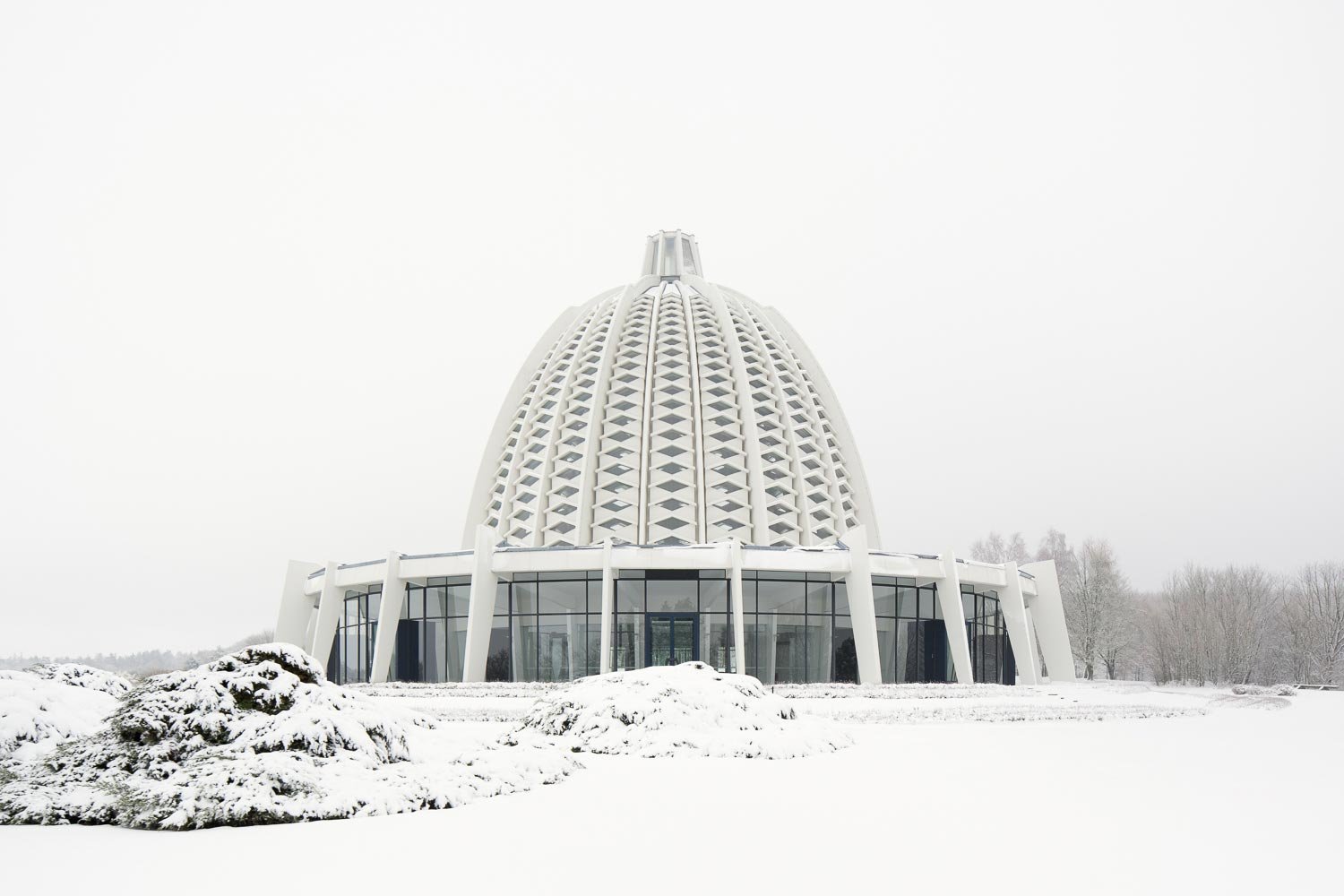 Zentralperspektive des europäischen Bahá'í-Tempels vor hellgrauem Himmel. Die lichtdurchlässige Kuppelarchitektur von Teuto Rocholl steht im Zentrum der schneebedeckten Parkanlage.