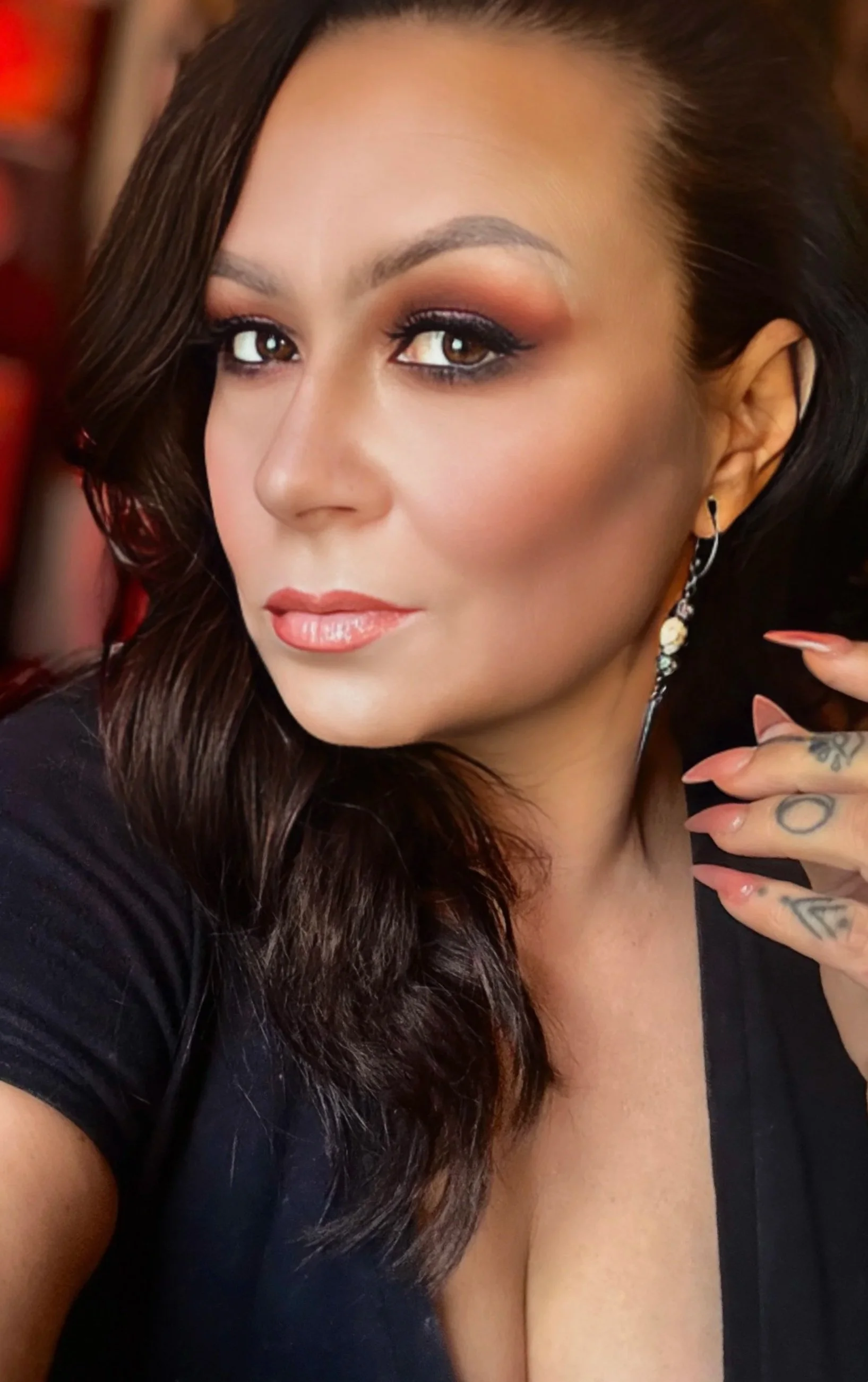 Close-up of a woman with long brown hair, makeup, and tattoos on her hand, wearing a black top, looking away from the camera.