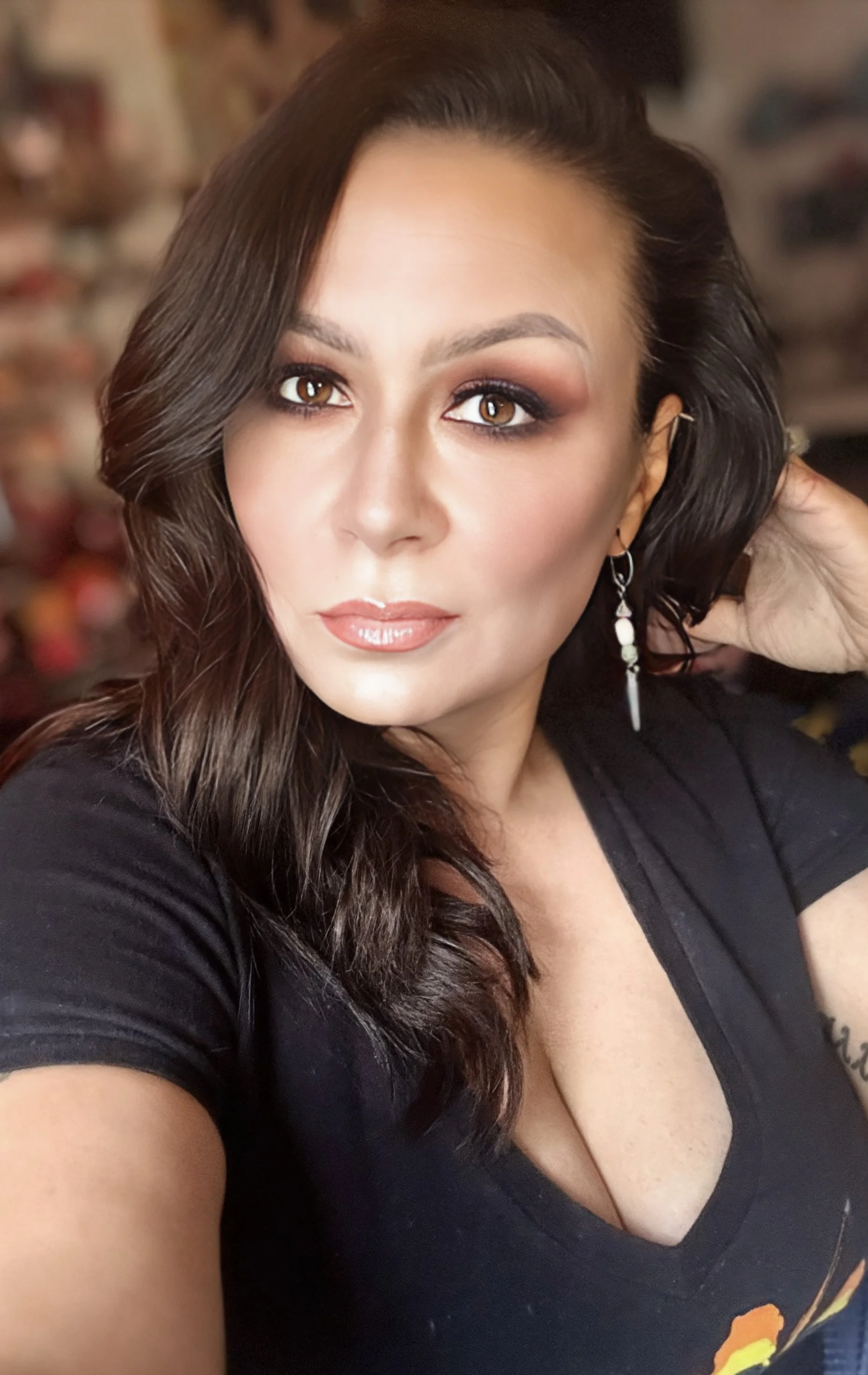 Close-up of a woman with long brown hair, wearing makeup, earrings, and a black top with a deep neckline, in a room with a blurred background.