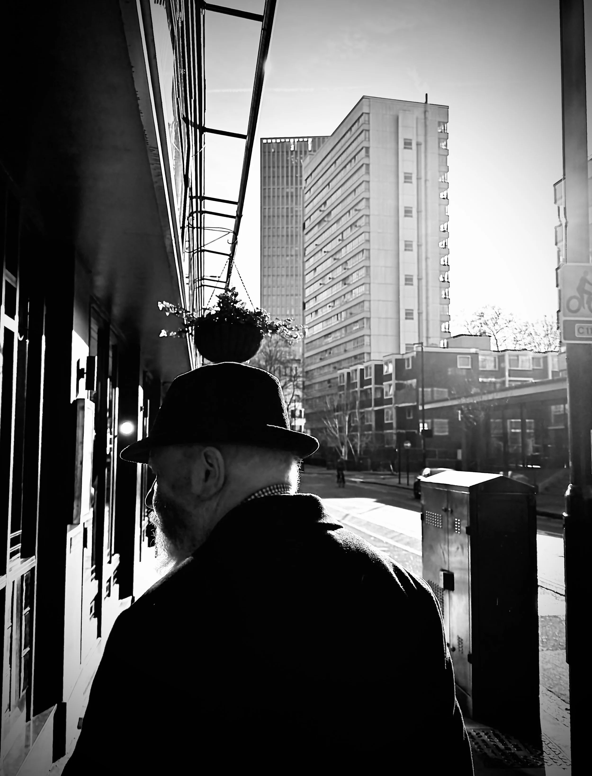 A black and white photo of an older man with a beard, wearing a fedora hat, seen from behind, walking on a city sidewalk during daytime. Tall buildings and a street with trees are visible in the background.