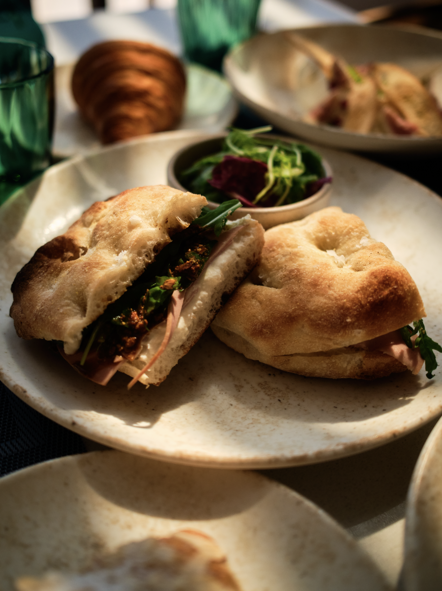 Close-up of a bread sandwich with greens and meat on a ceramic plate, with a side of salad and glasses in the background.