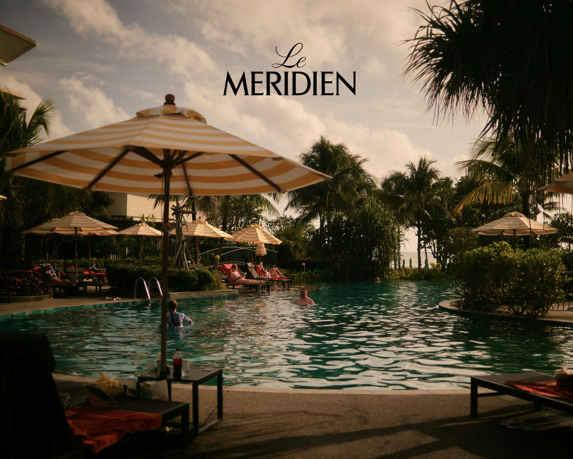 People swimming and relaxing around a pool at Le Meridien hotel, with palm trees and umbrellas providing shade on a sunny day.