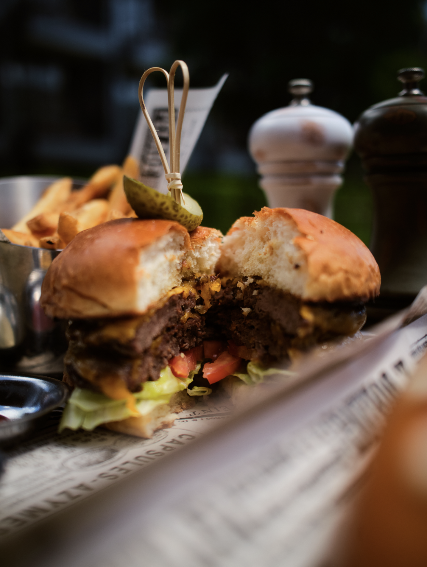 Close-up of half-eaten burger with lettuce, tomato, cheese, and cooked meat patty, topped with a pickle and served with French fries in the background.