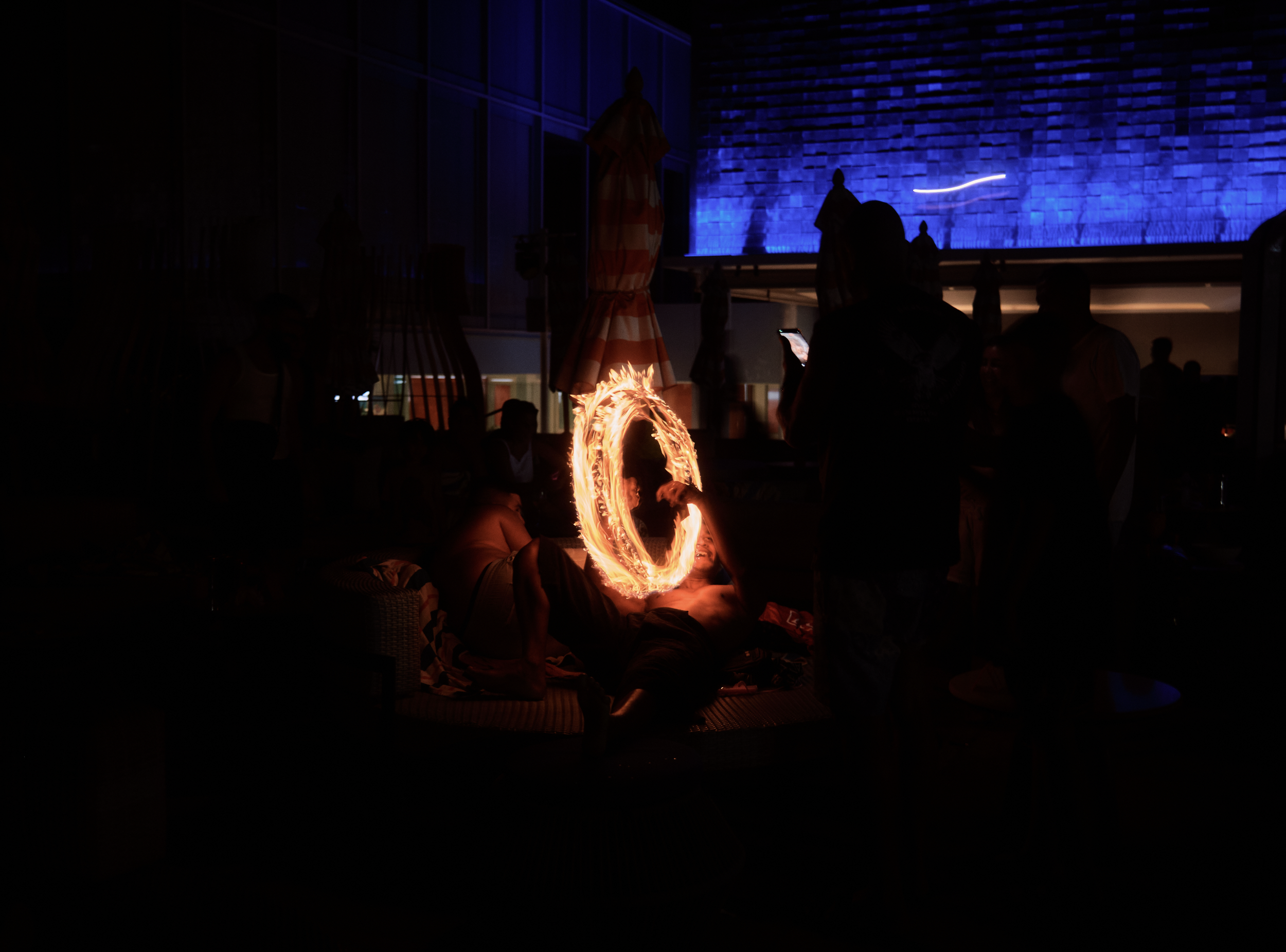 People gathered in a darkened room watching a fire performance with flames creating a bright circular shape, illuminated by blue lighting and a large blue-lit wall in the background.