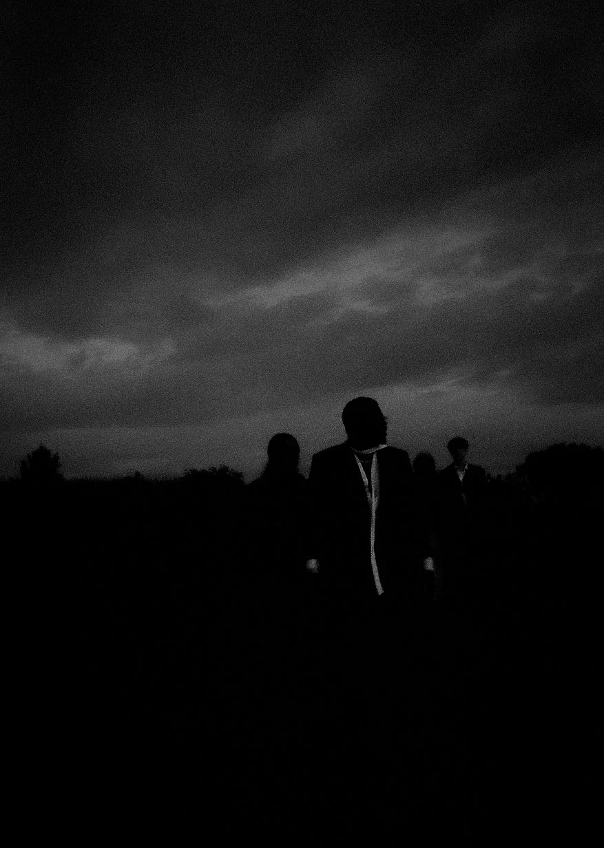 Silhouettes of four people walking outdoors at dusk with a dark, cloudy sky overhead.