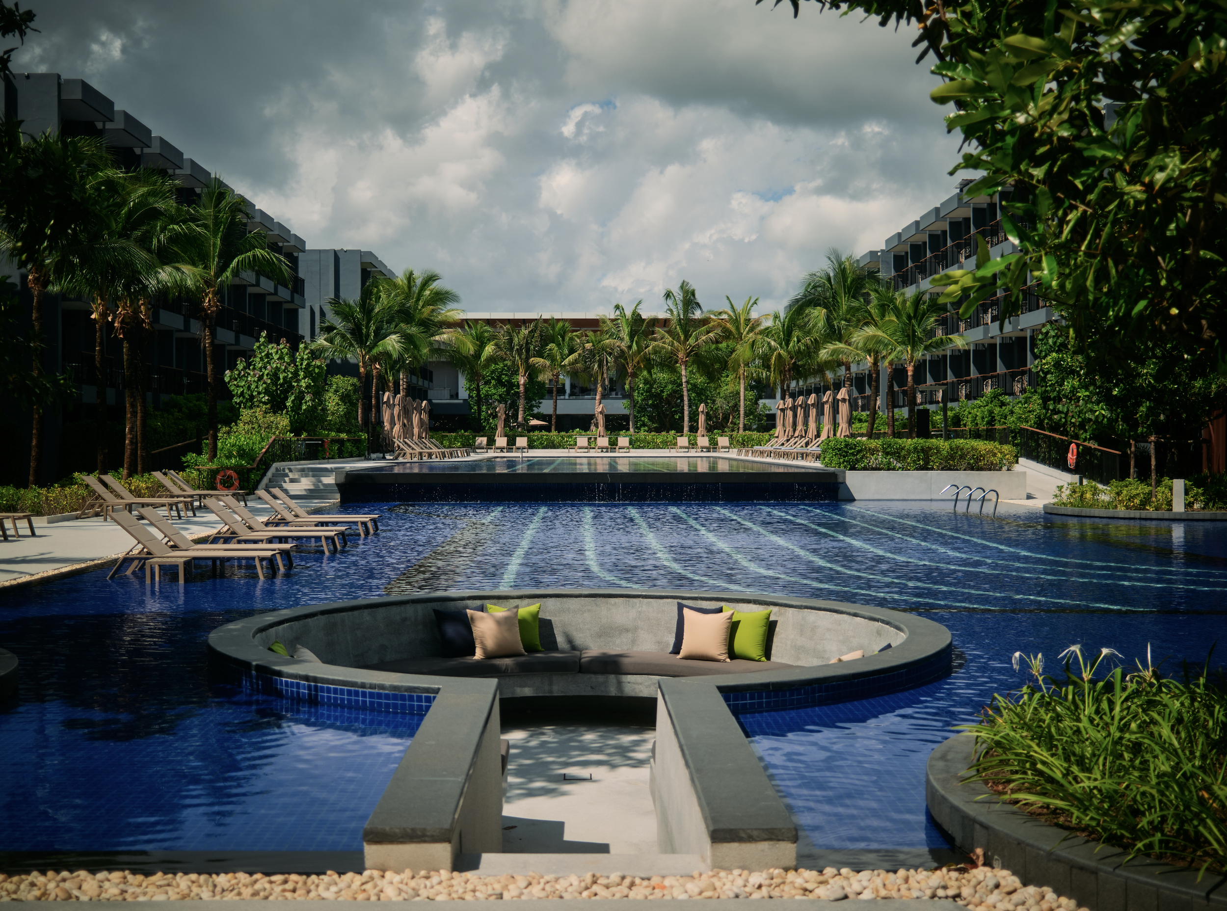 A luxurious outdoor swimming pool area with a hot tub in the foreground, lounge chairs, umbrellas, lush green palm trees and shrubbery, multi-story modern buildings, and a cloudy sky in the background.