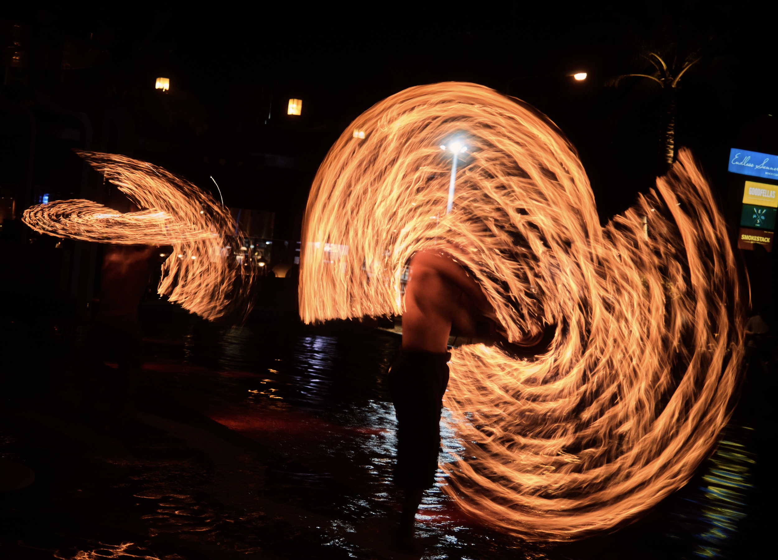 Fiery circle formations created by performers spinning fire poi at night, with people silhouetted against the bright flames. Anemoia.