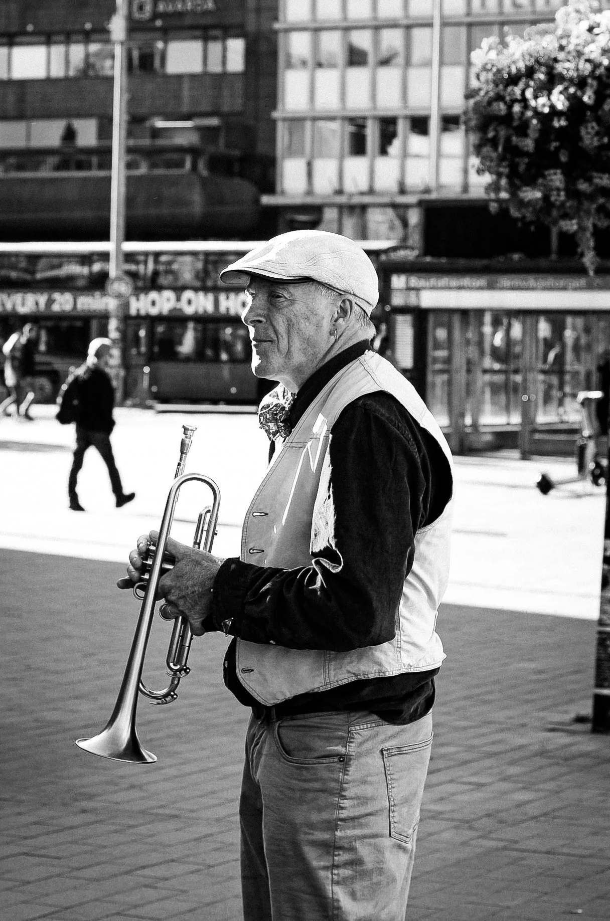 A man with a hat and a bowtie holding a trumpet on a city street, with pedestrians and buildings in the background.