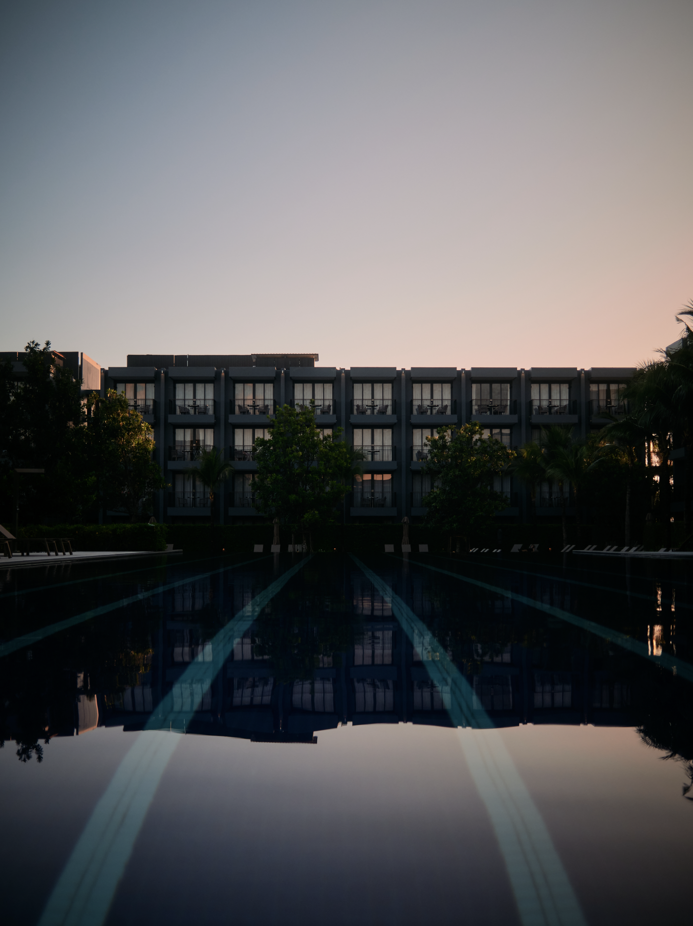A multi-story hotel or apartment building with balconies at sunset. Trees surround the building, and a swimming pool with blue lines reflects the building and sky in the foreground.