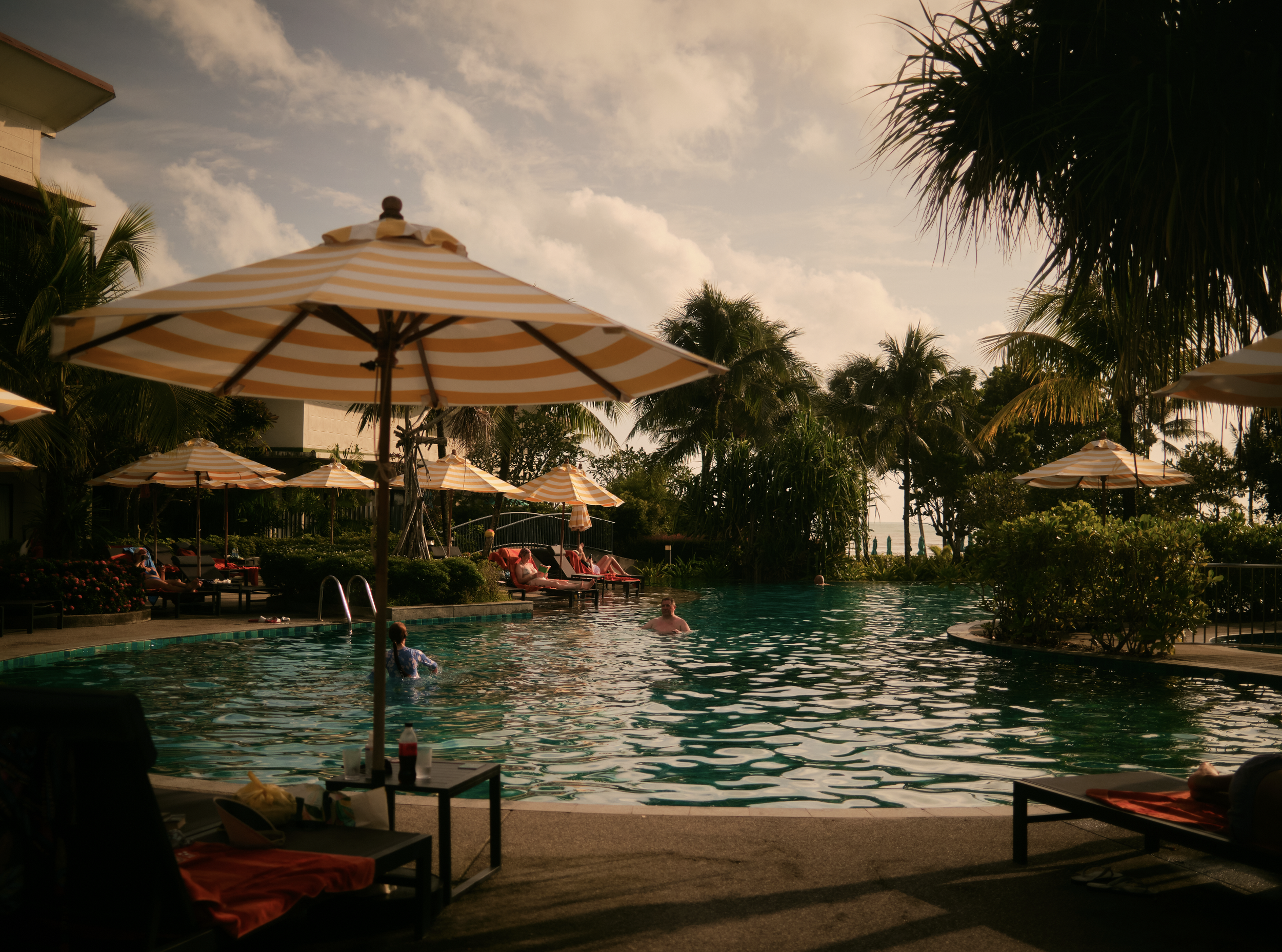 A tropical resort swimming pool area with yellow and white striped umbrellas, palm trees, and lounge chairs, during the daytime with a partly cloudy sky. Anemoia.