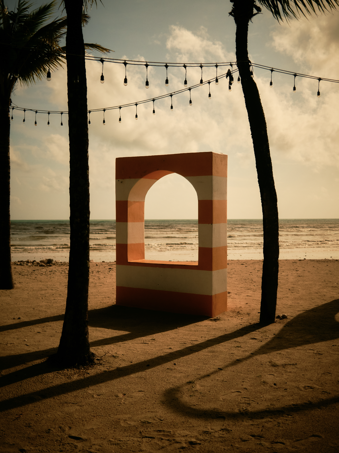 Beach scene at sunset with a striped pink and white arch, palm trees, and string lights hanging above the sand and ocean in the background.