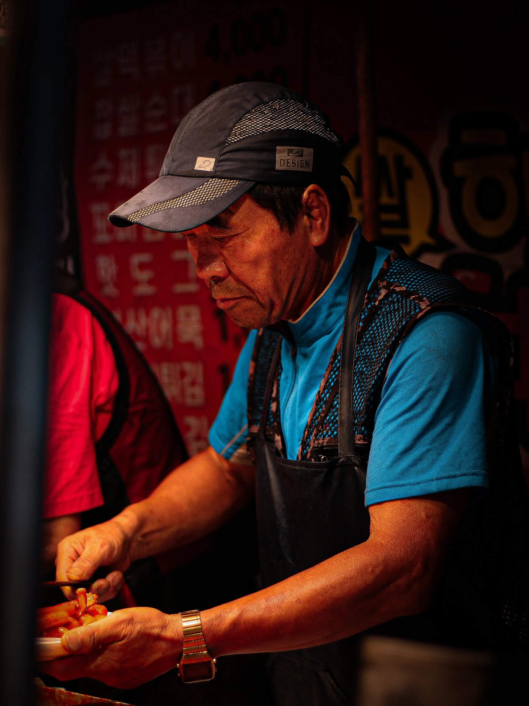 An older man wearing a black skull cap, blue shirt, and black apron, preparing food in a dimly lit kitchen or food stall with red and black signage in the background.