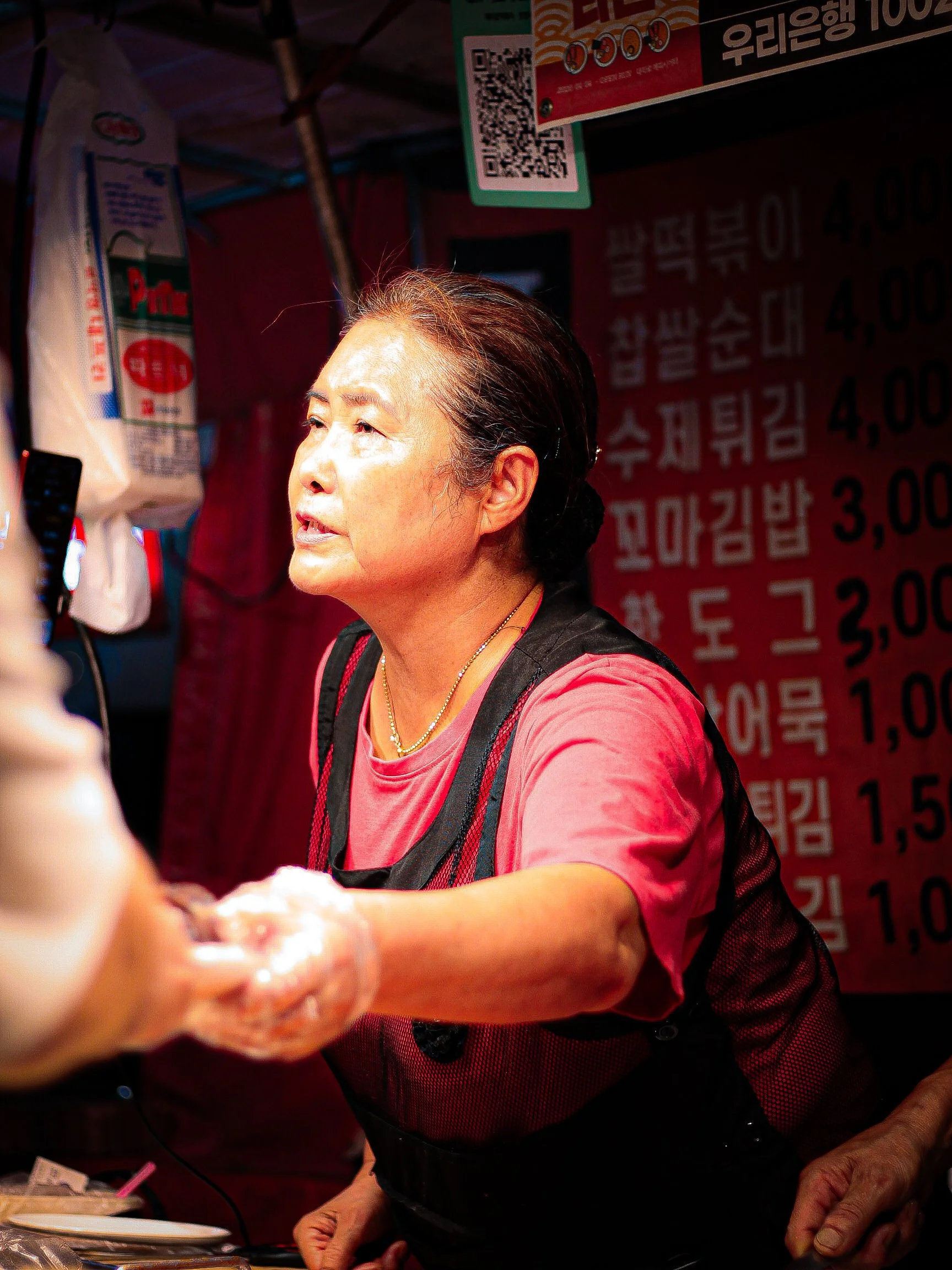A woman working at a food stall, wearing a pink shirt and black apron, with Korean menu and prices in the background.