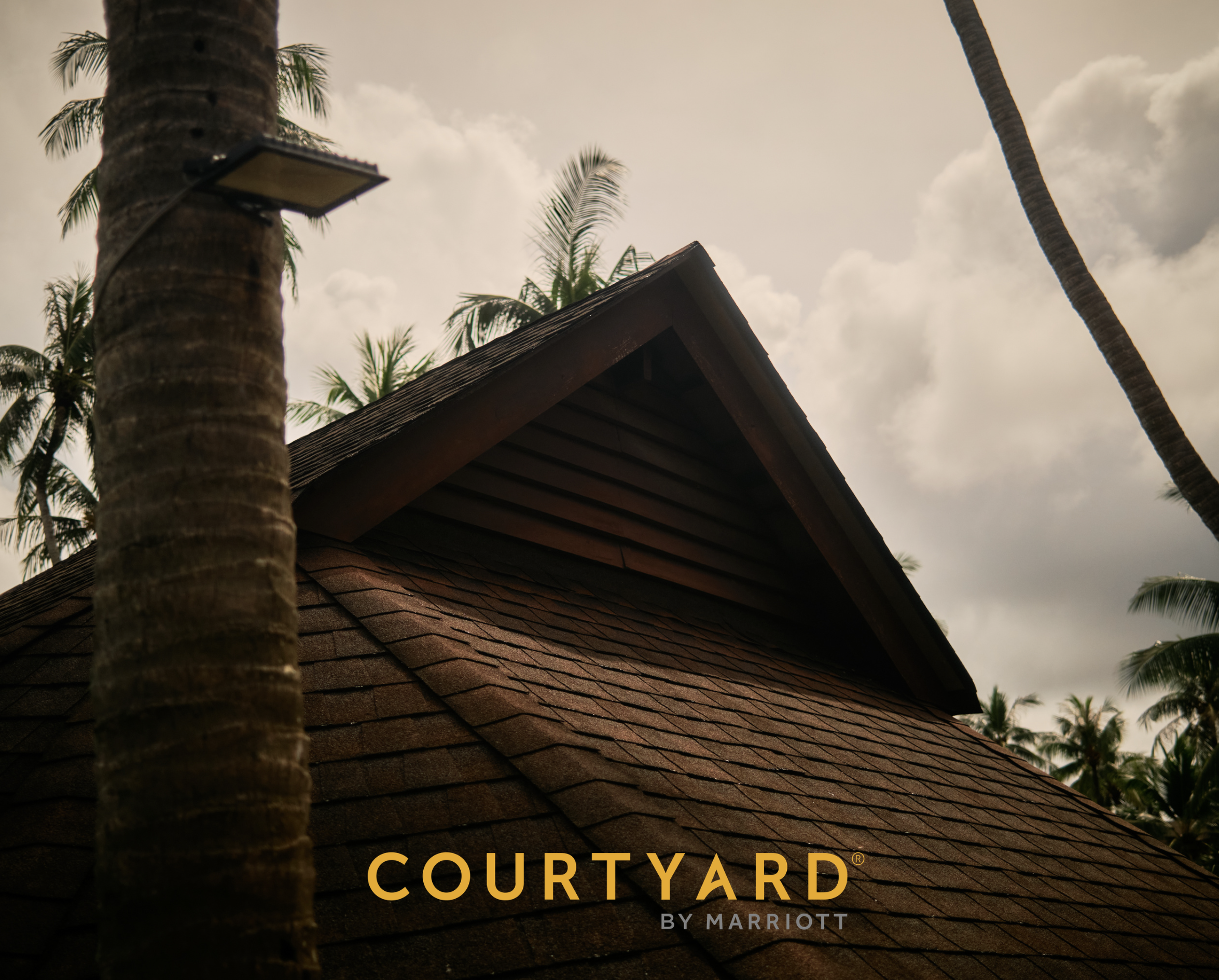 Close-up of a wooden rooftop with palm trees and a partly cloudy sky in the background. The text 'Countryard by Marriott' is visible at the bottom of the image.