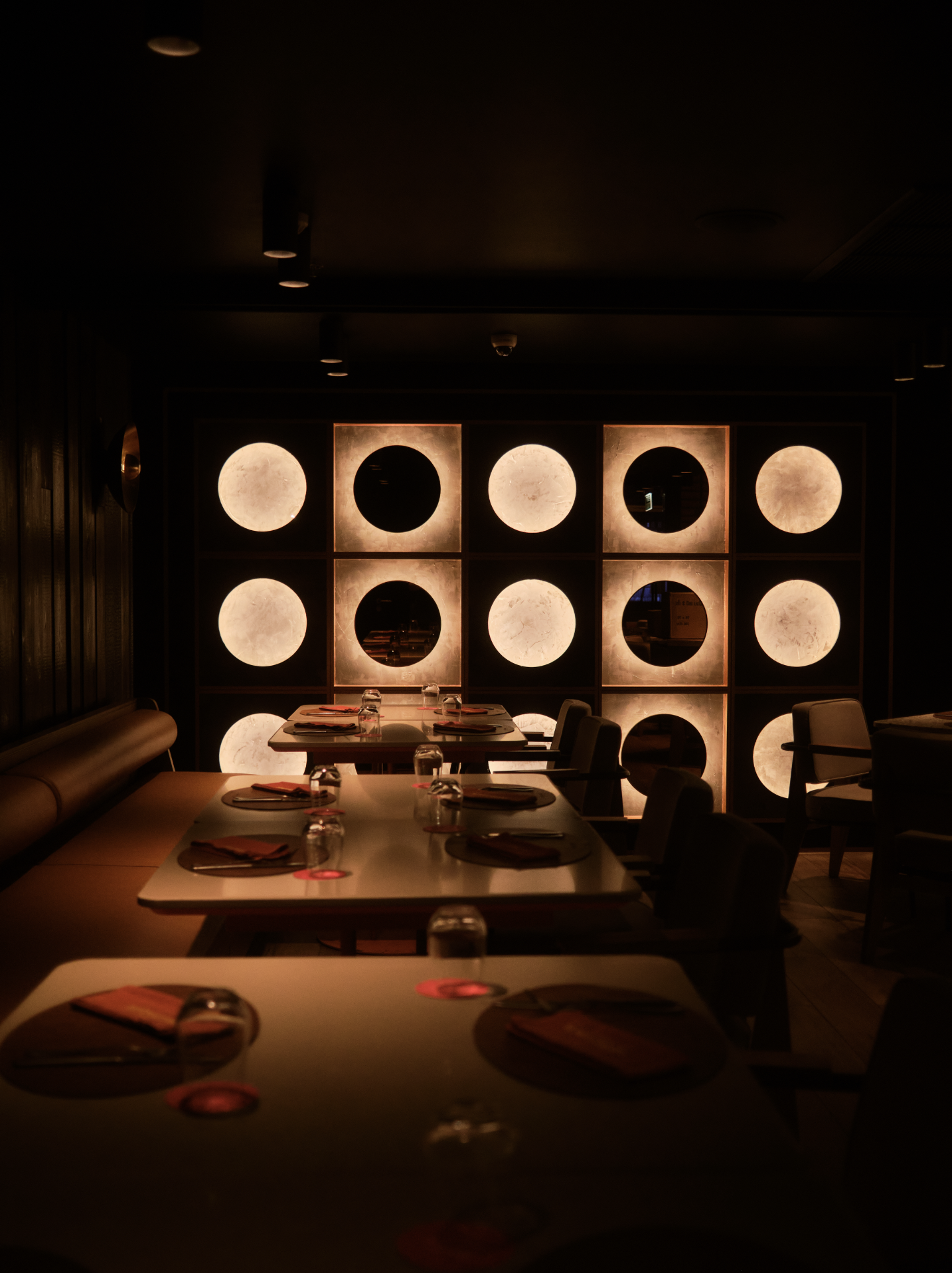 Dimly lit restaurant interior with tables set for dining, featuring cutlery, napkins, and glasses, and a decorative illuminated wall with circular and square panels. Anemoia.