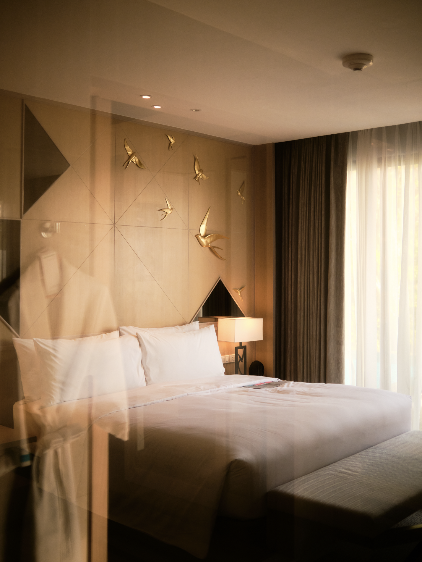 A hotel room with a large bed, a bedside lamp, and a decorative wall featuring gold hummingbird sculptures and geometric panels, with a window covered by dark curtains. Anemoia.