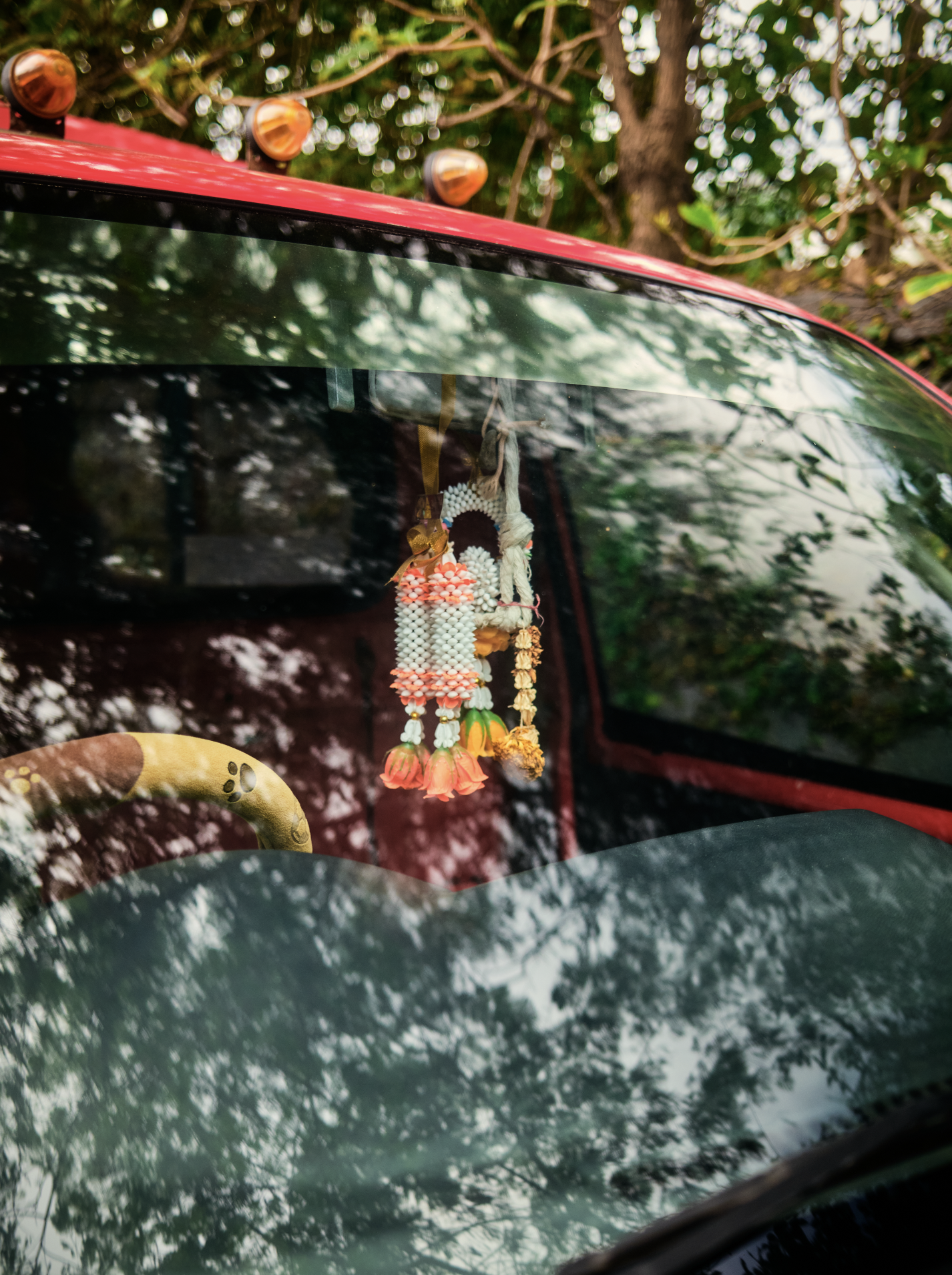 Close-up of the front windshield of a red vehicle with decorative hanging ornaments inside, trees and foliage reflected on the glass. Anemoia.