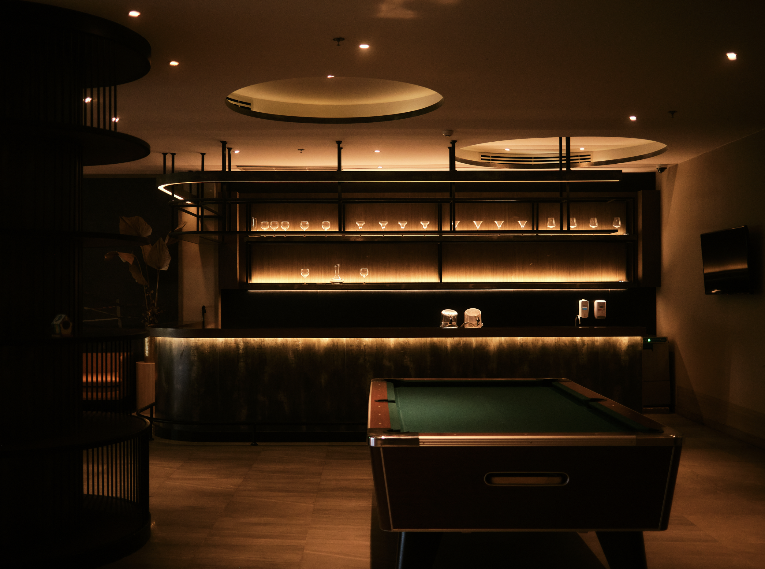 Dimly lit bar with a row of glasses on shelves behind the bar counter, a pool table in the foreground, and round ceiling fixtures with embedded lights.