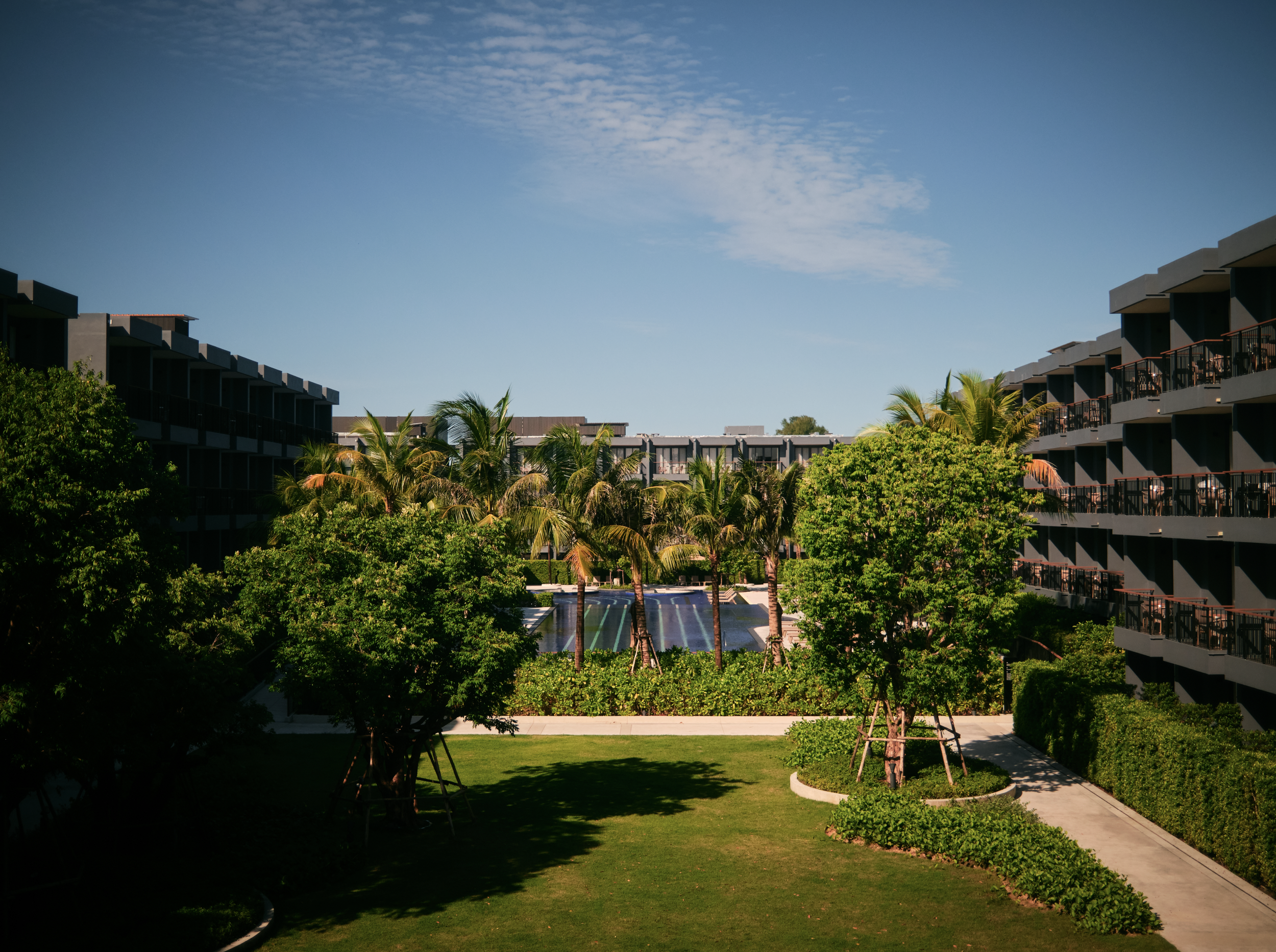 View of a modern apartment complex with multiple stories, balconies, and glass railings, surrounded by lush green trees and a well-maintained lawn under a bright blue sky with some clouds.