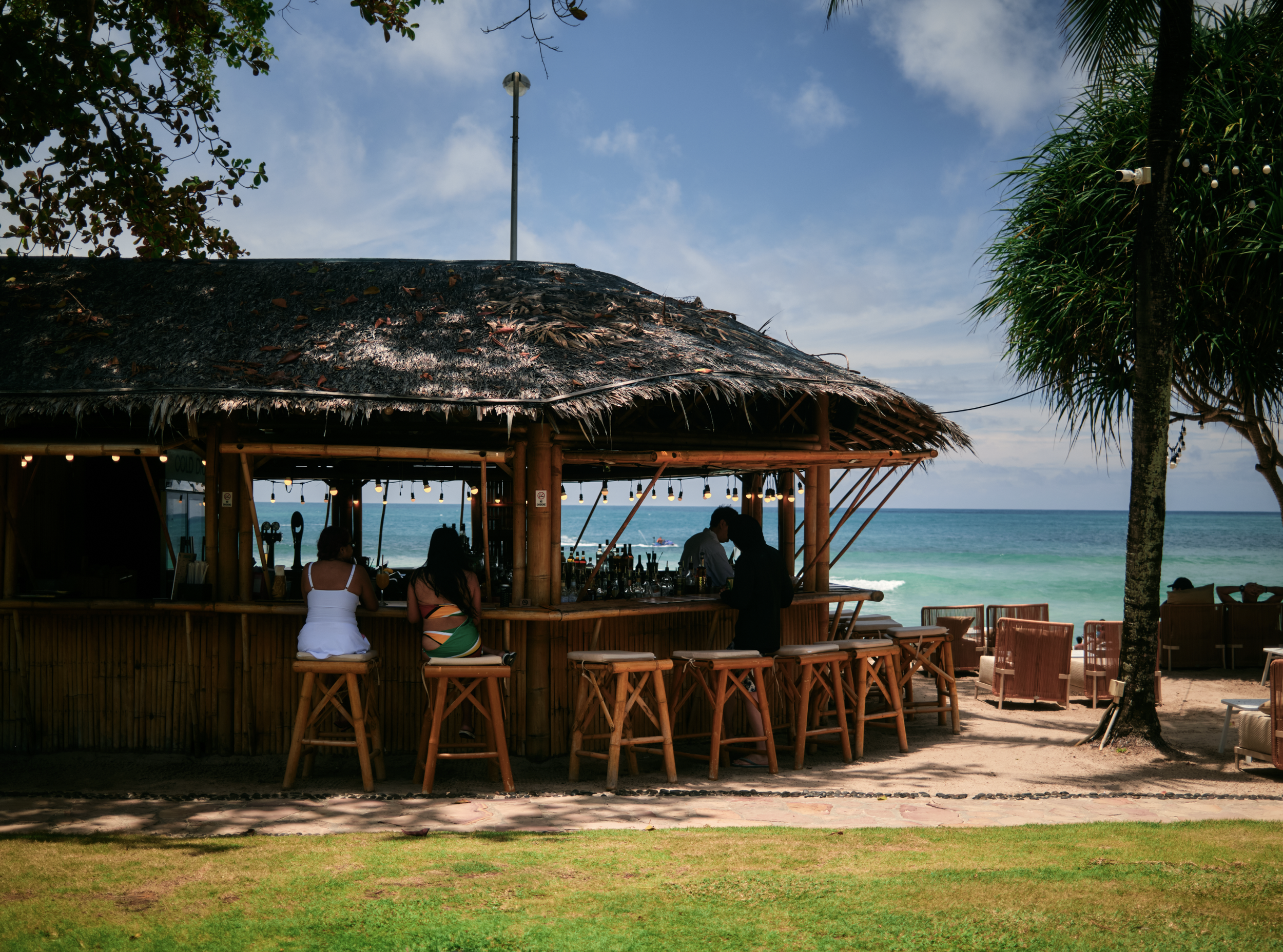 Sunset at a beachside bar with thatched roof, bar stools, and people sitting inside, overlooking the ocean with a clear sky.