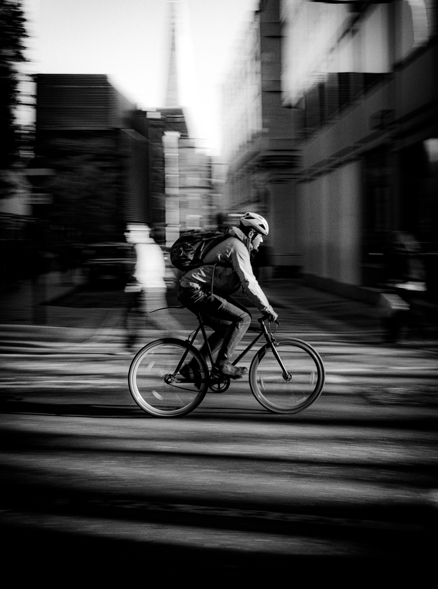 A person riding a bicycle on a city street during daytime, with tall buildings in the background and appearing to be moving quickly as the photo is blurred.