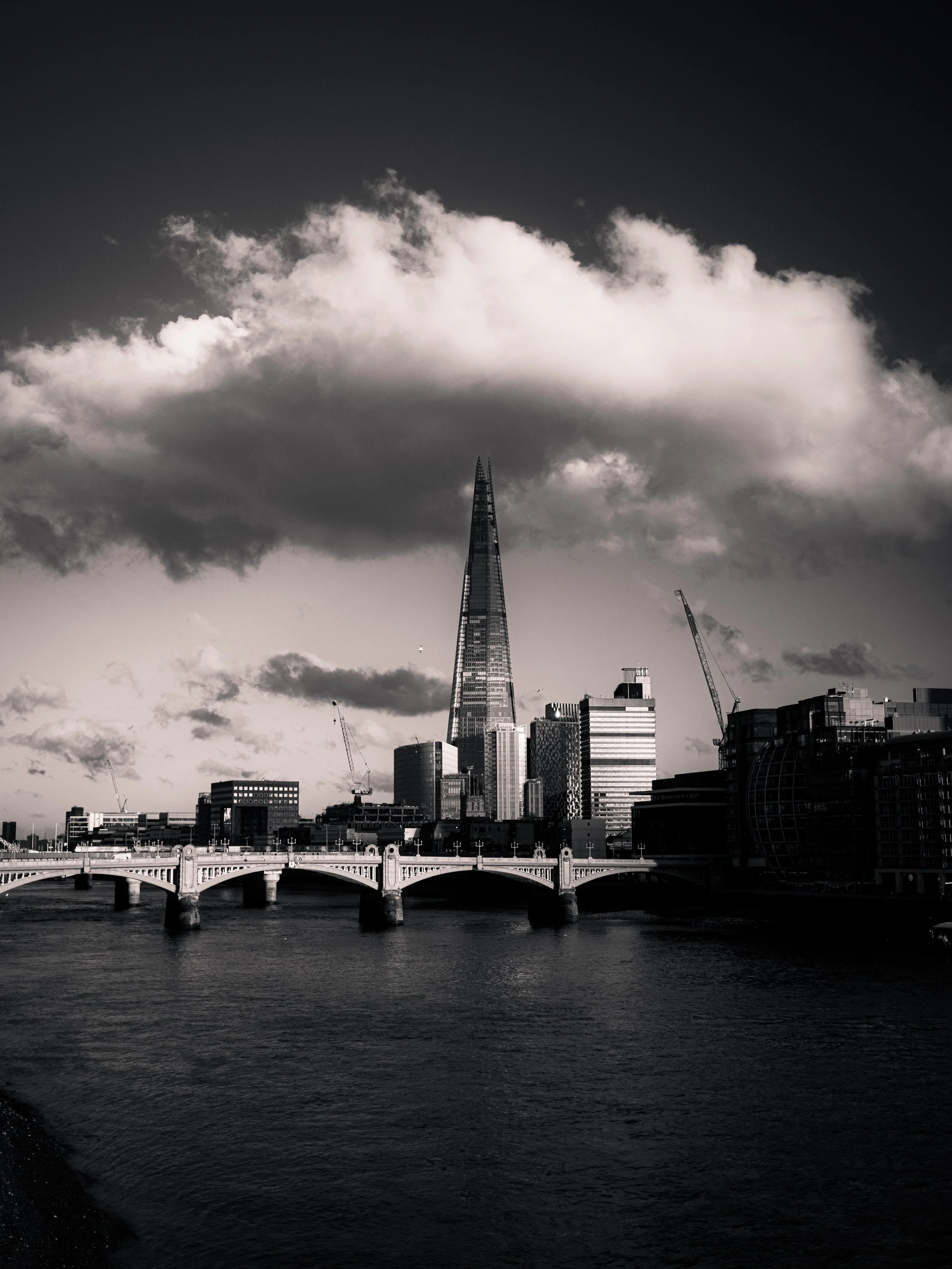 Black and white photo of a city skyline with tall skyscrapers, a bridge over a river in the foreground, and a large cloud in the sky.