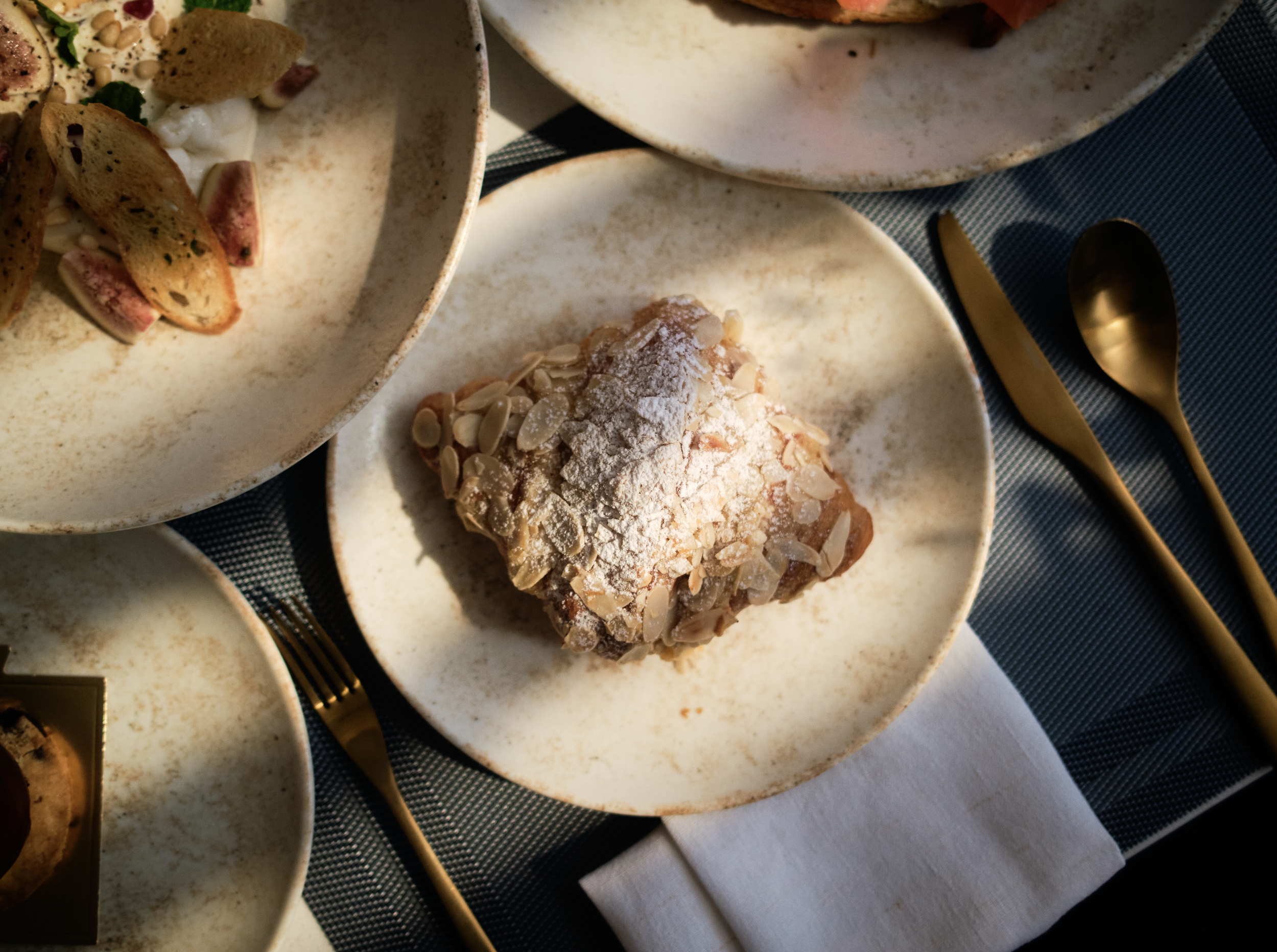 Close-up of almond-covered pastry on a plate with gold-colored cutlery on a dark blue cloth.