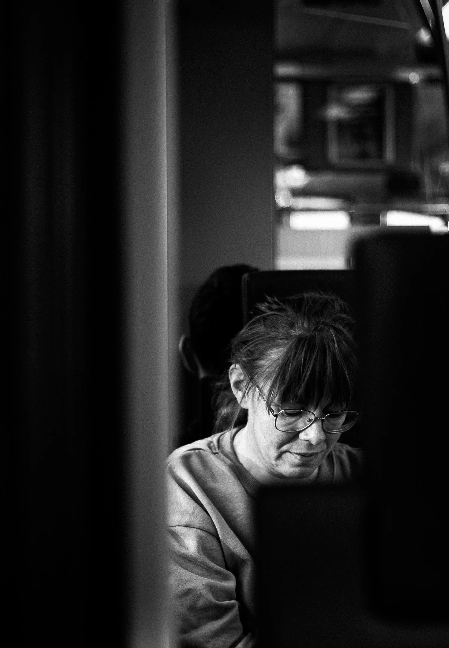 A woman with glasses working on a computer, viewed through a narrow gap or space, in a black and white photo.