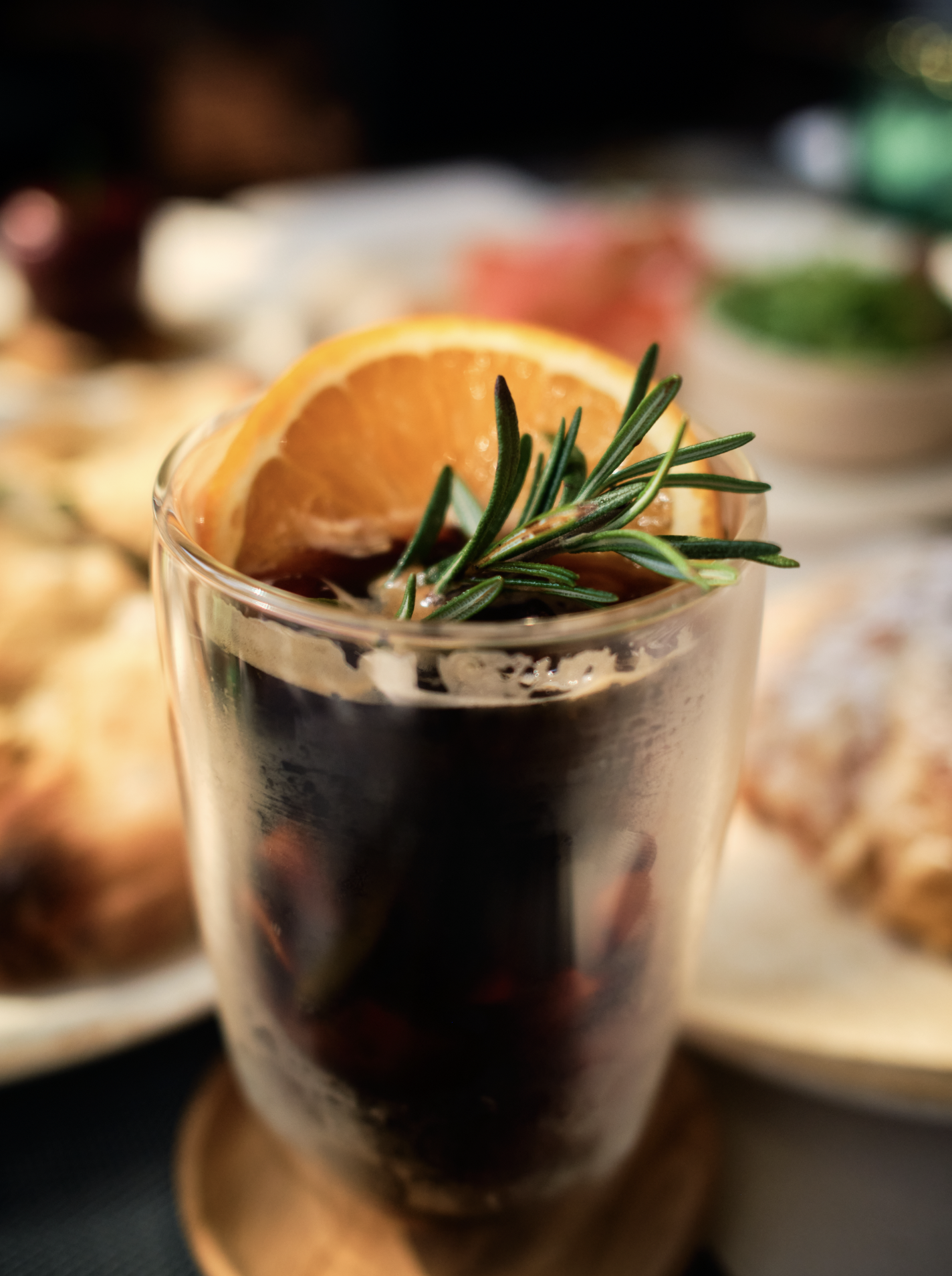 A glass of dark beverage garnished with an orange slice and a sprig of rosemary, with blurred background of other drinks and snacks.