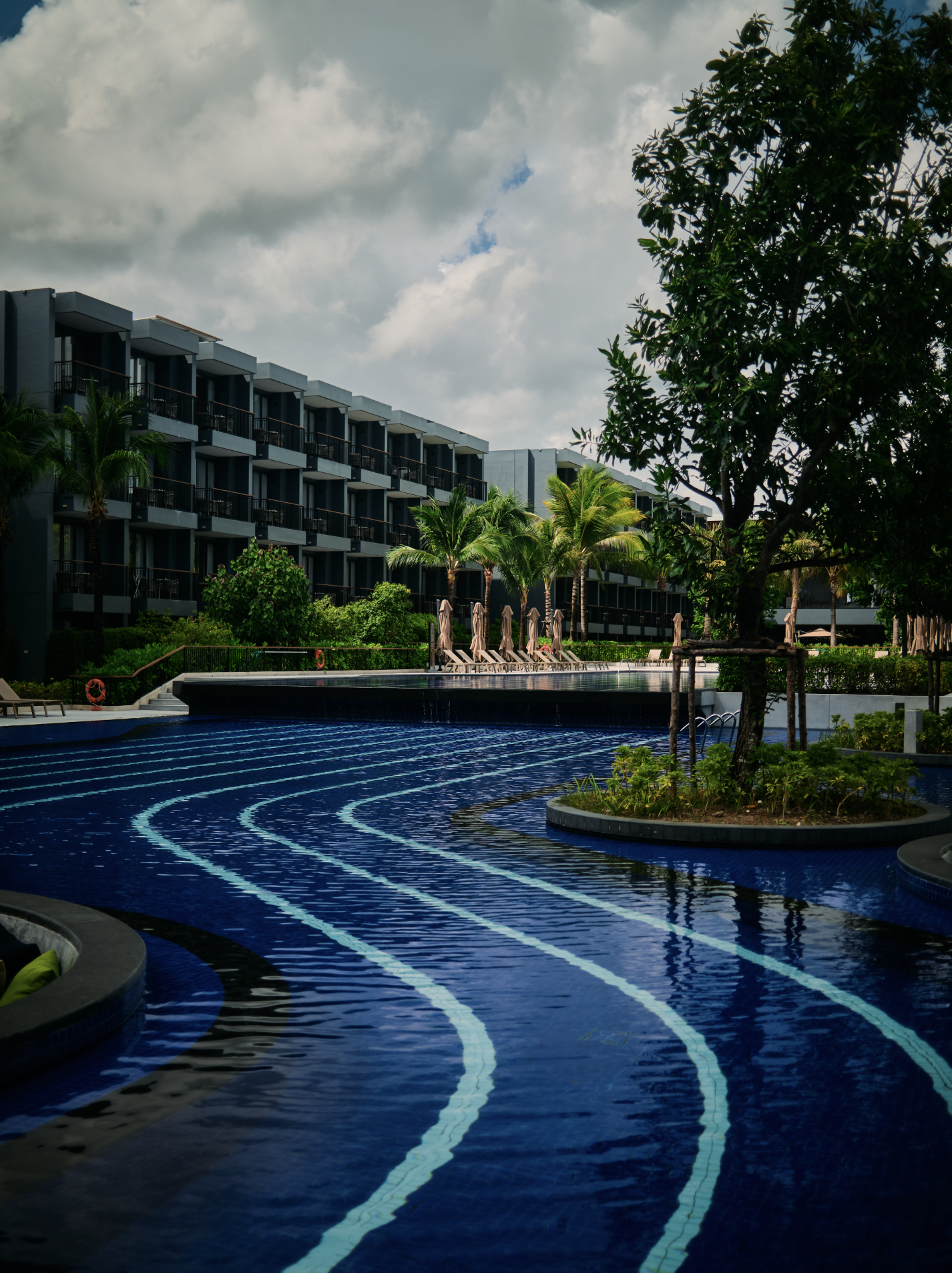 A modern hotel with a swimming pool surrounded by lounge chairs and lush greenery, including palm trees, under a partly cloudy sky.