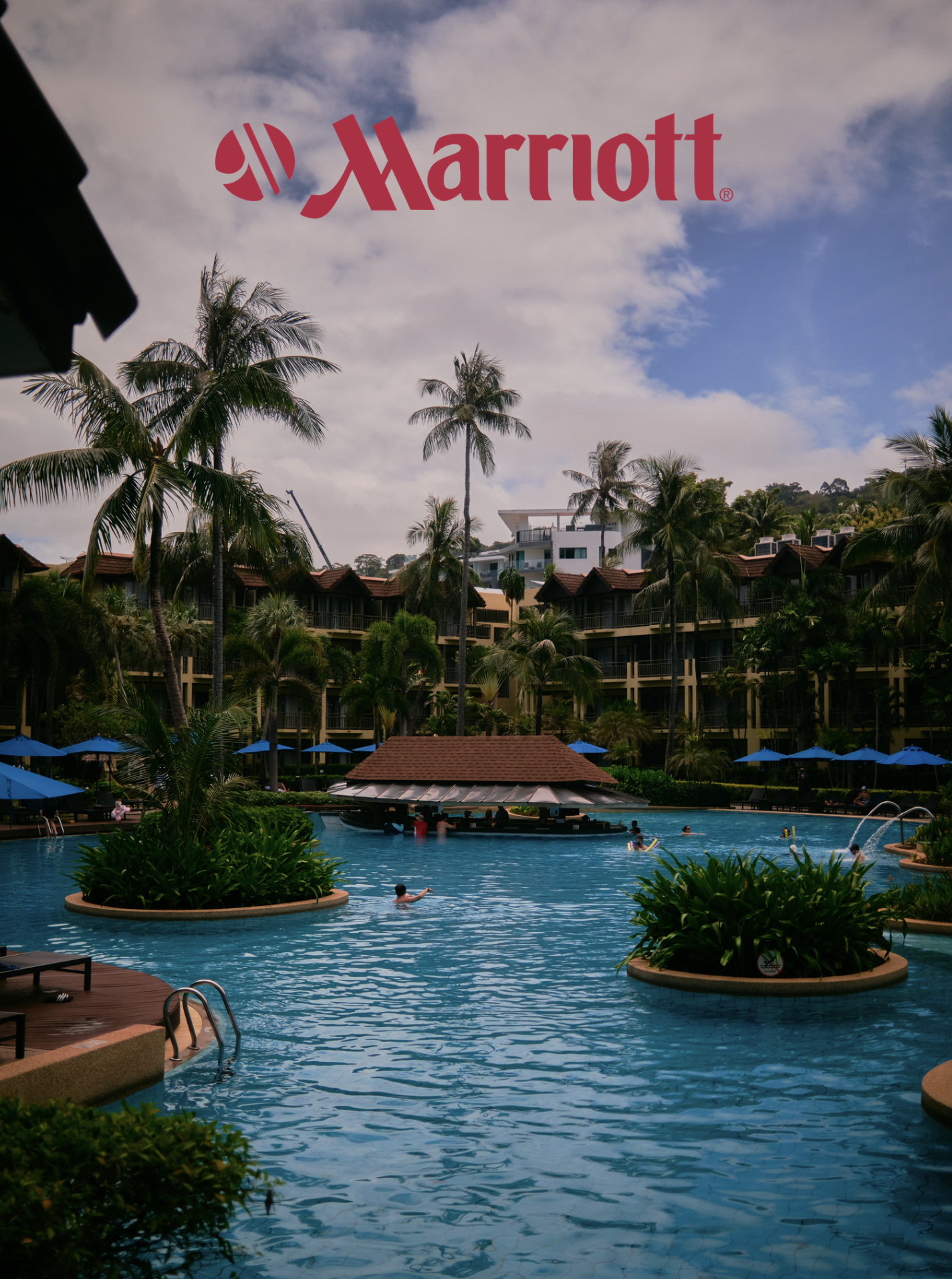 A swimming pool at a Marriott hotel with palm trees, umbrellas, and a poolside bar, surrounded by hotel rooms and lush greenery under a partly cloudy sky.