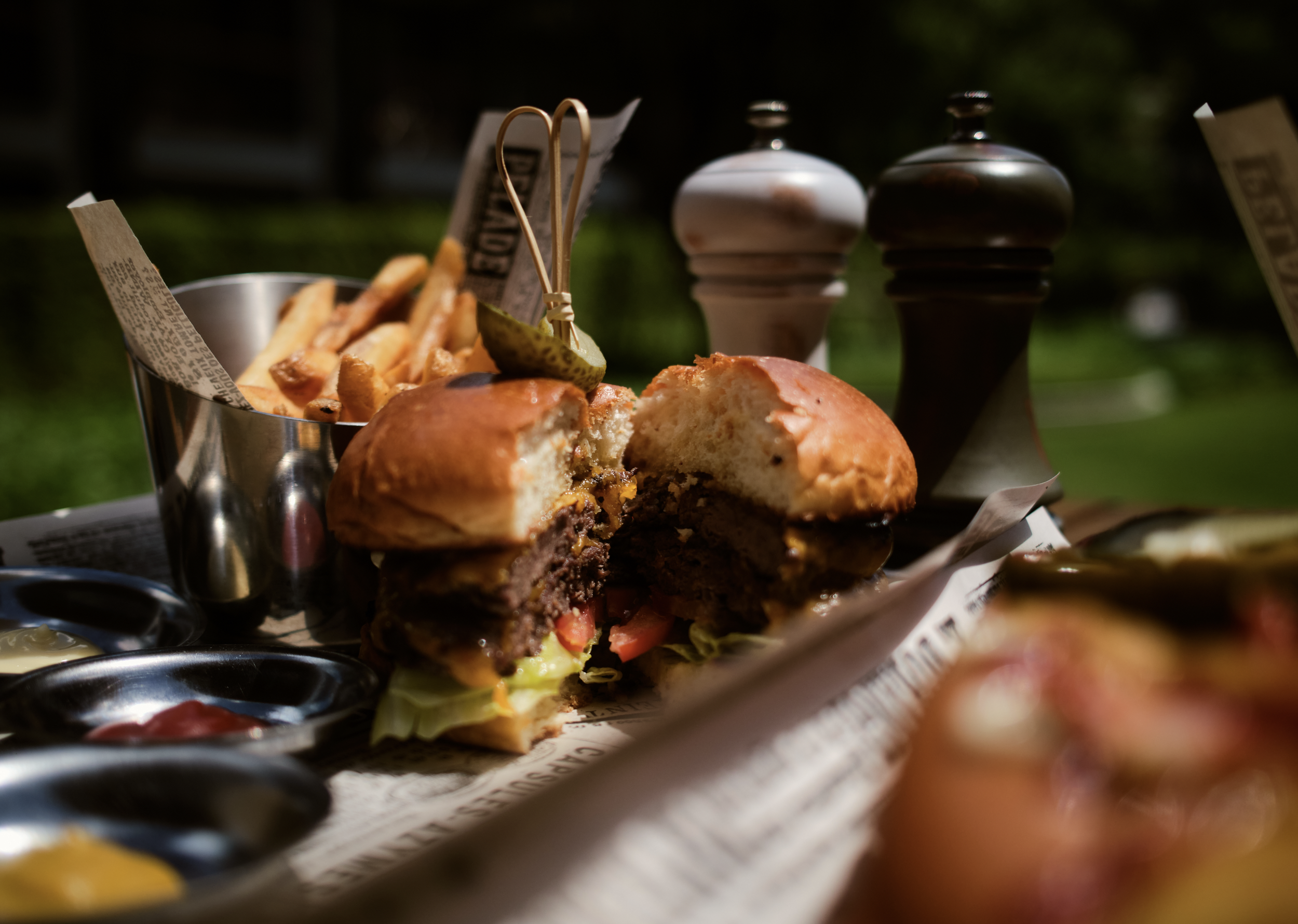 A burger in a bun with visible lettuce, tomato, and meat patty, along with a side of French fries, and condiments in small containers, on a metal tray with parchment paper, outdoors with a dark background.